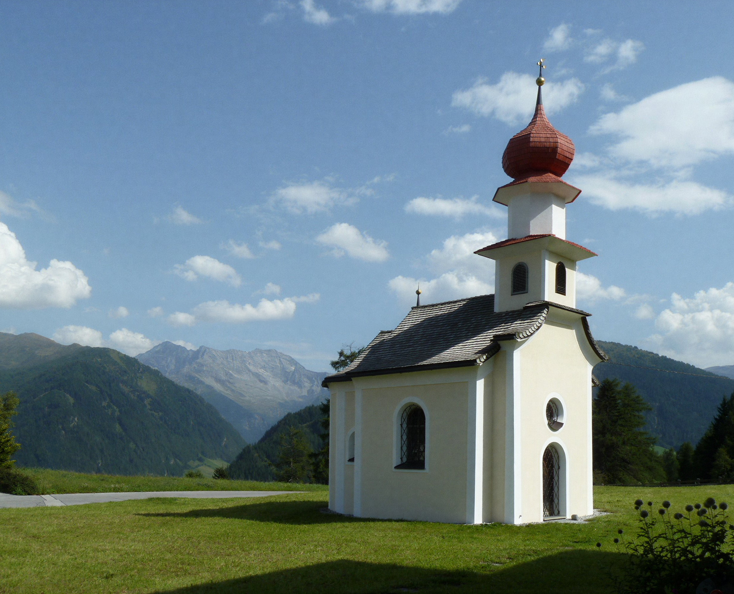 Kleine weiße Kirche mit rotem Dach in einer grünen Landschaft mit Bergen im Hintergrund und einem blauen Himmel mit Wolken.