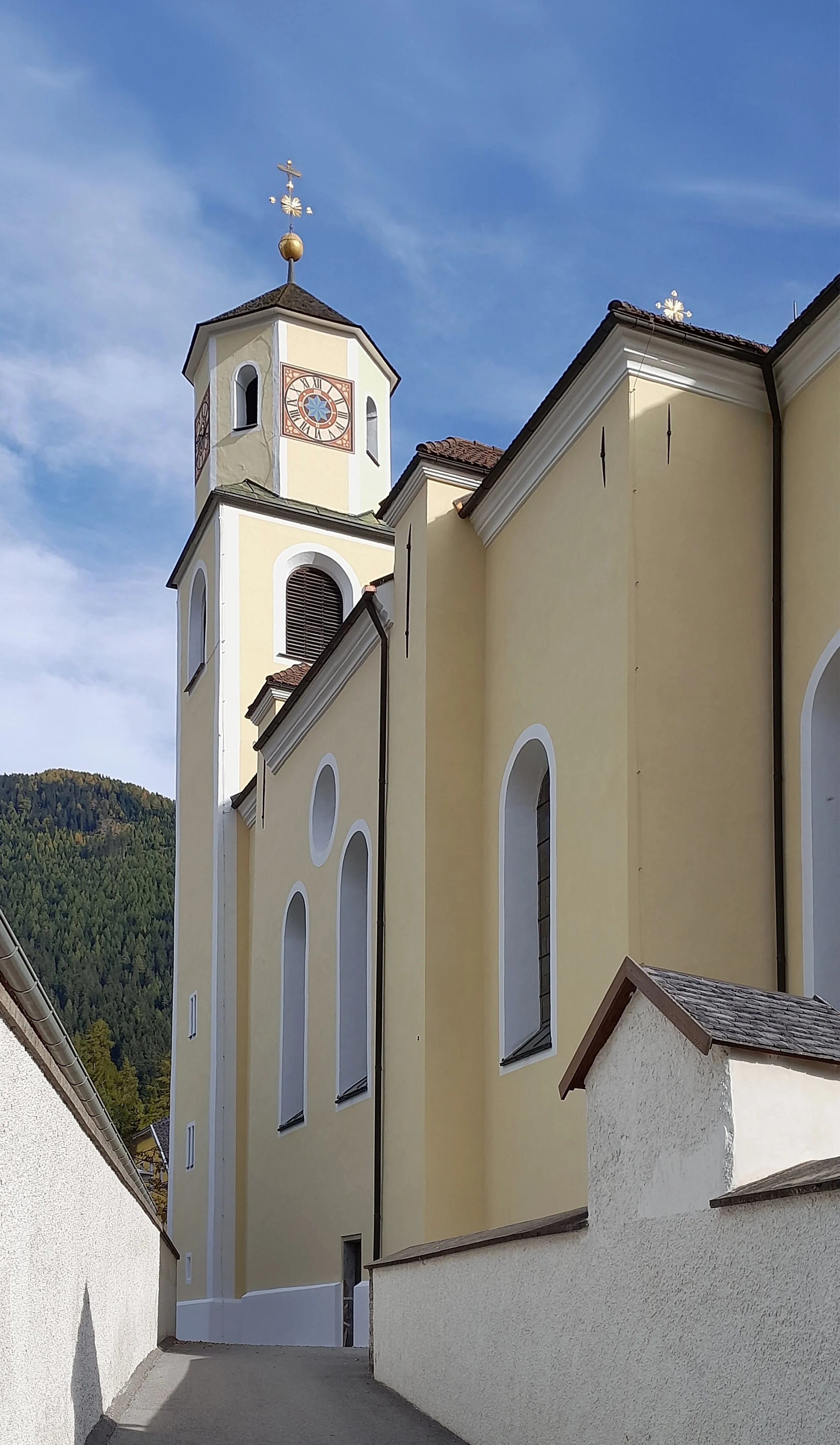 Die Frontansicht einer Kirche mit gelben Wänden, hohen schmalen Fenstern und einem Uhrturm mit einer Wetterfahne unter klarem Himmel.