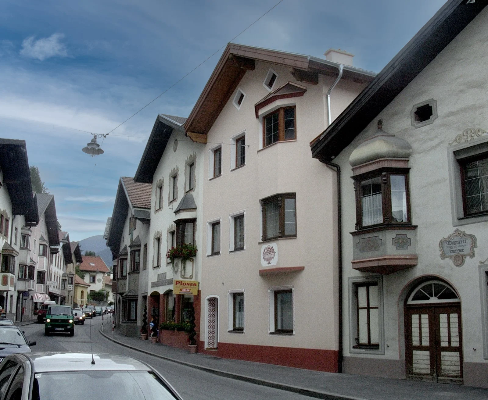 Traditionelle deutsche Häuser mit Sprossenfenstern und Verzierungen an der Fassade, eine Straße mit geparkten Autos und Berge im Hintergrund.