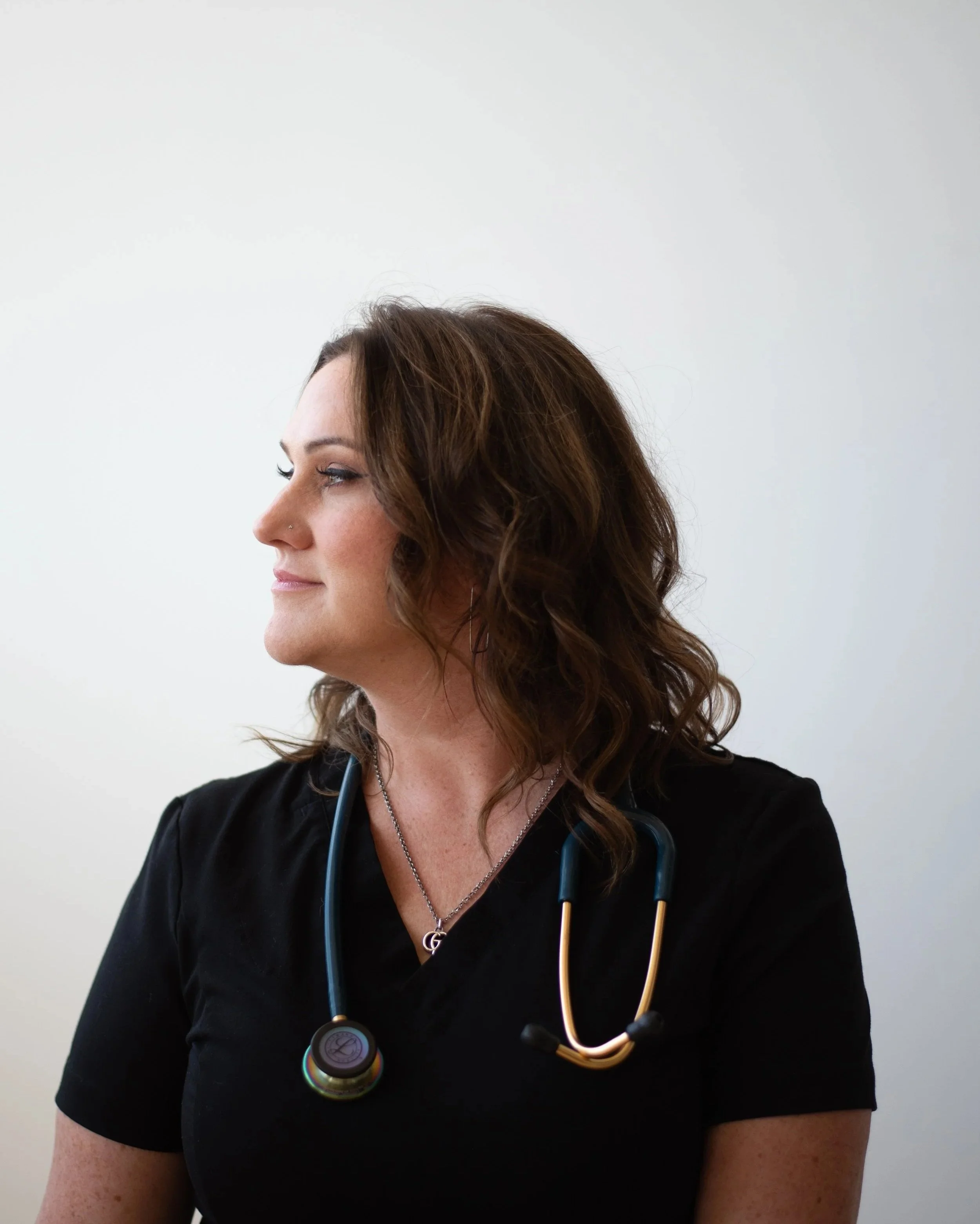 Side profile of a female healthcare professional wearing a black uniform and a stethoscope around her neck against a plain gray background.