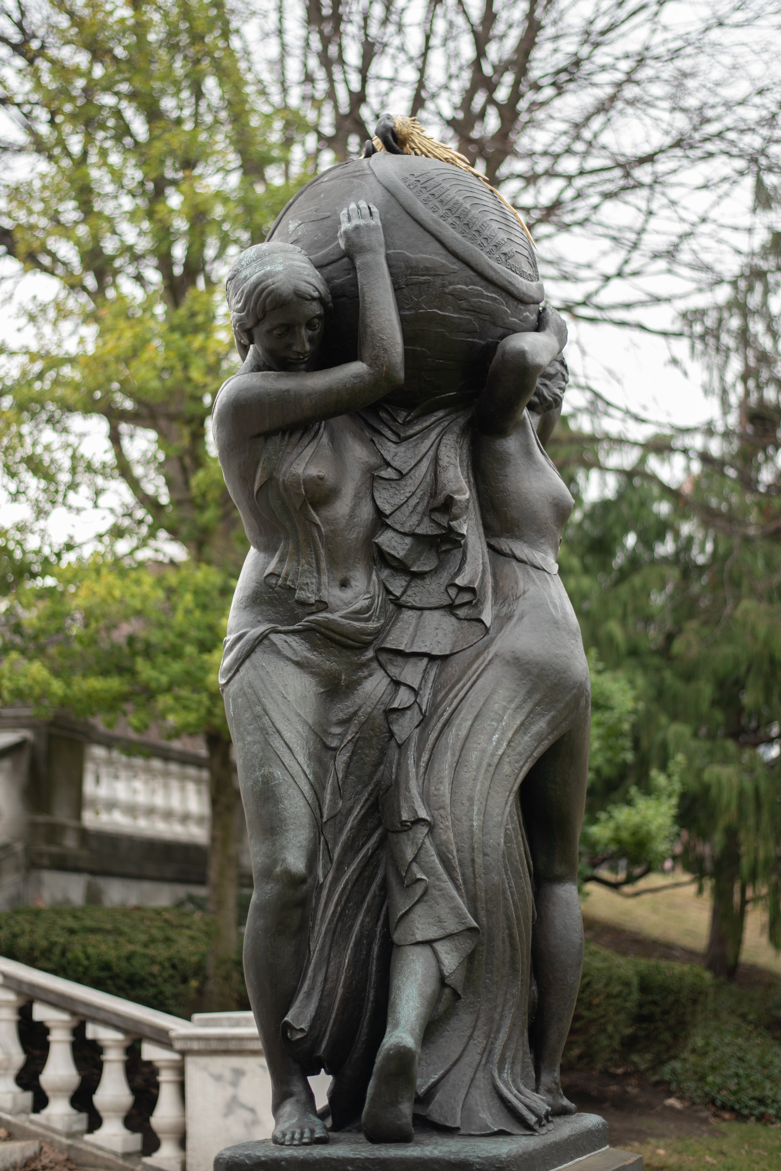 Statue of a woman carrying a large globe on her shoulders with a bird perched on top. The woman has long flowing hair and is dressed in a draped garment, standing outdoors with trees in the background.
