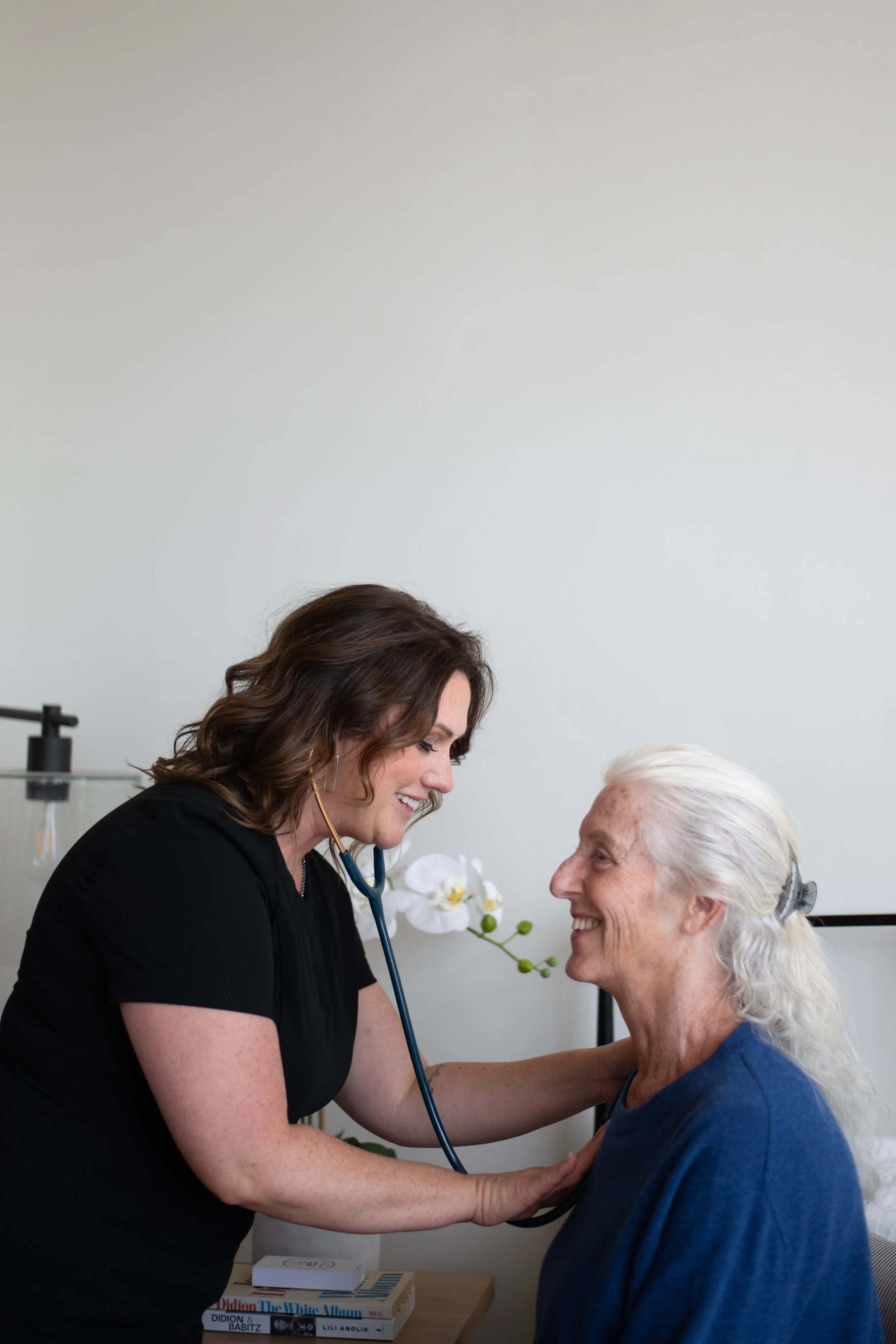 A healthcare professional, a woman with dark hair, smiling, using a stethoscope to listen to an elderly woman's chest, who has white hair, sitting and smiling, inside a medical office with a white wall and a potted orchid plant in the background.