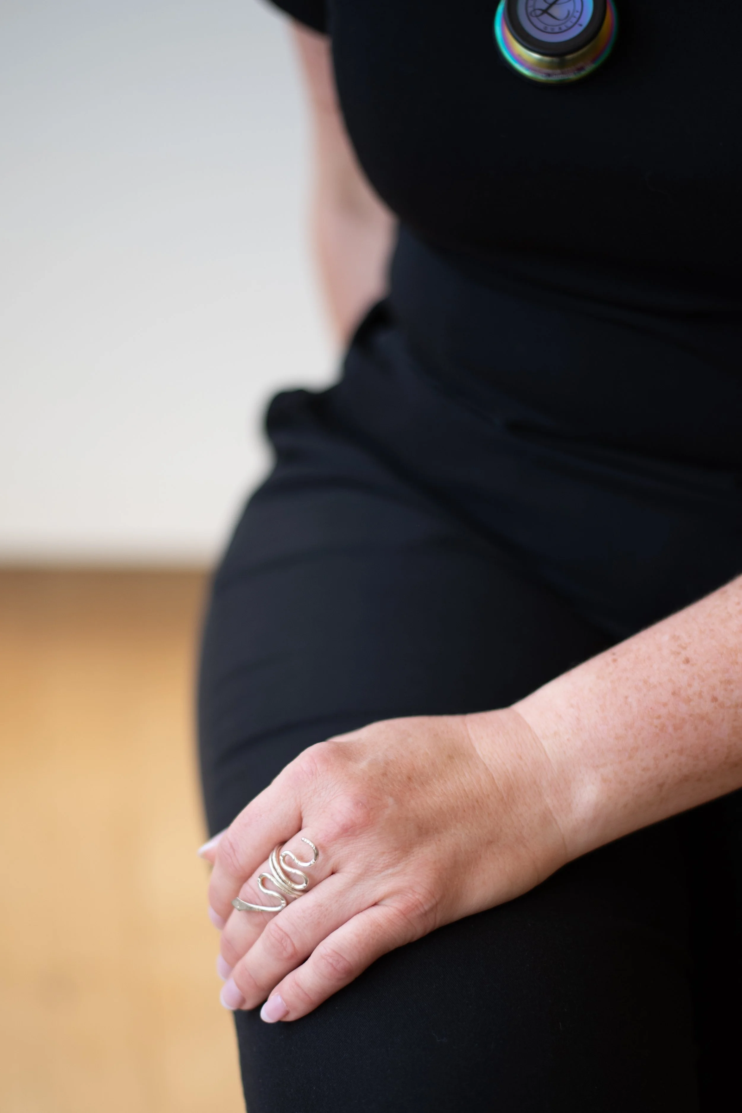 Close-up of a person's hand resting on their lap, wearing a silver ring with the word 'SUS' on it, and a black shirt or dress. The background is blurry and indicates an indoor setting.