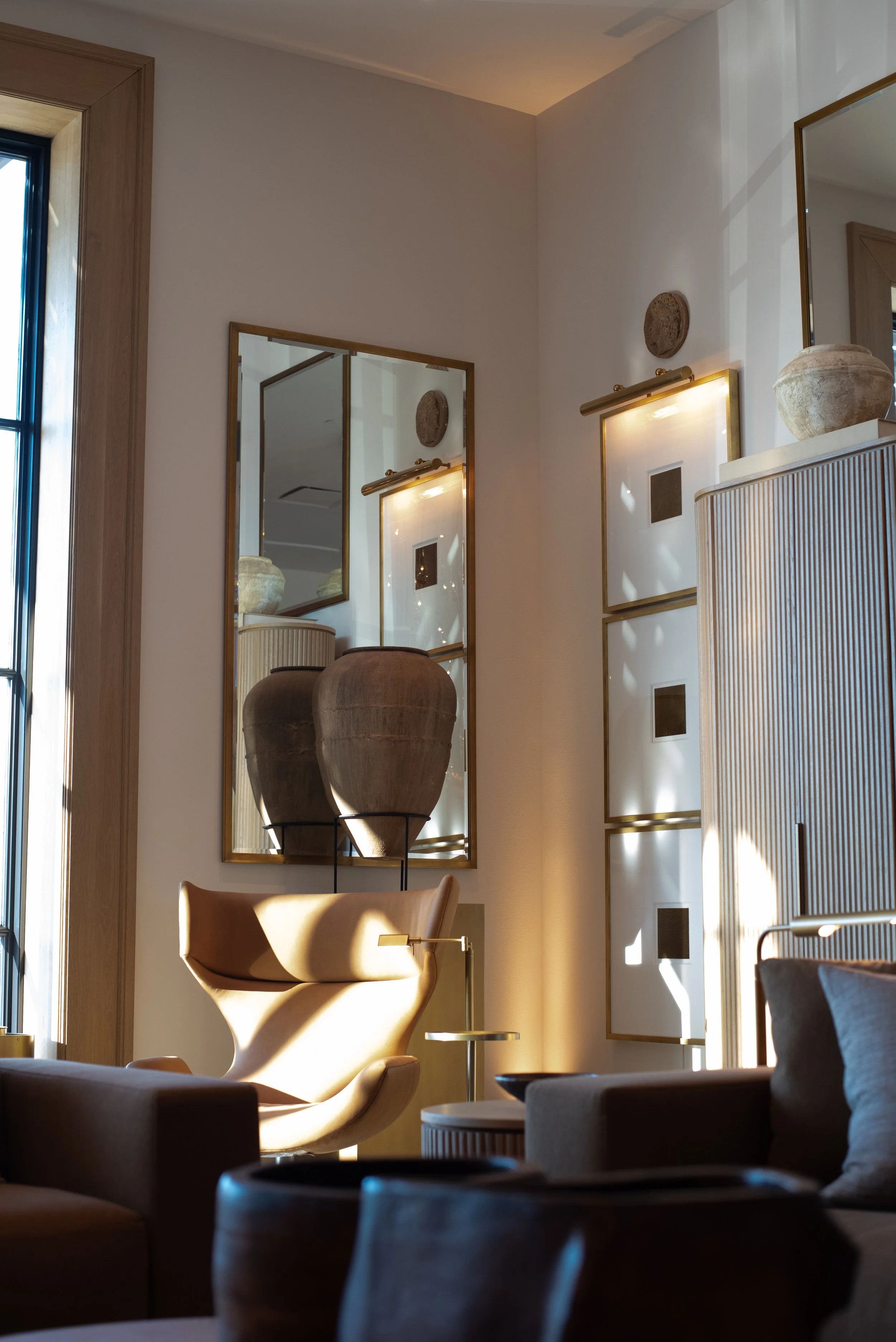 A living room with a beige armchair, potted plant, and wall mirrors reflected in the windows. Sunlight filters through the window, casting shadows on the wall and furniture.