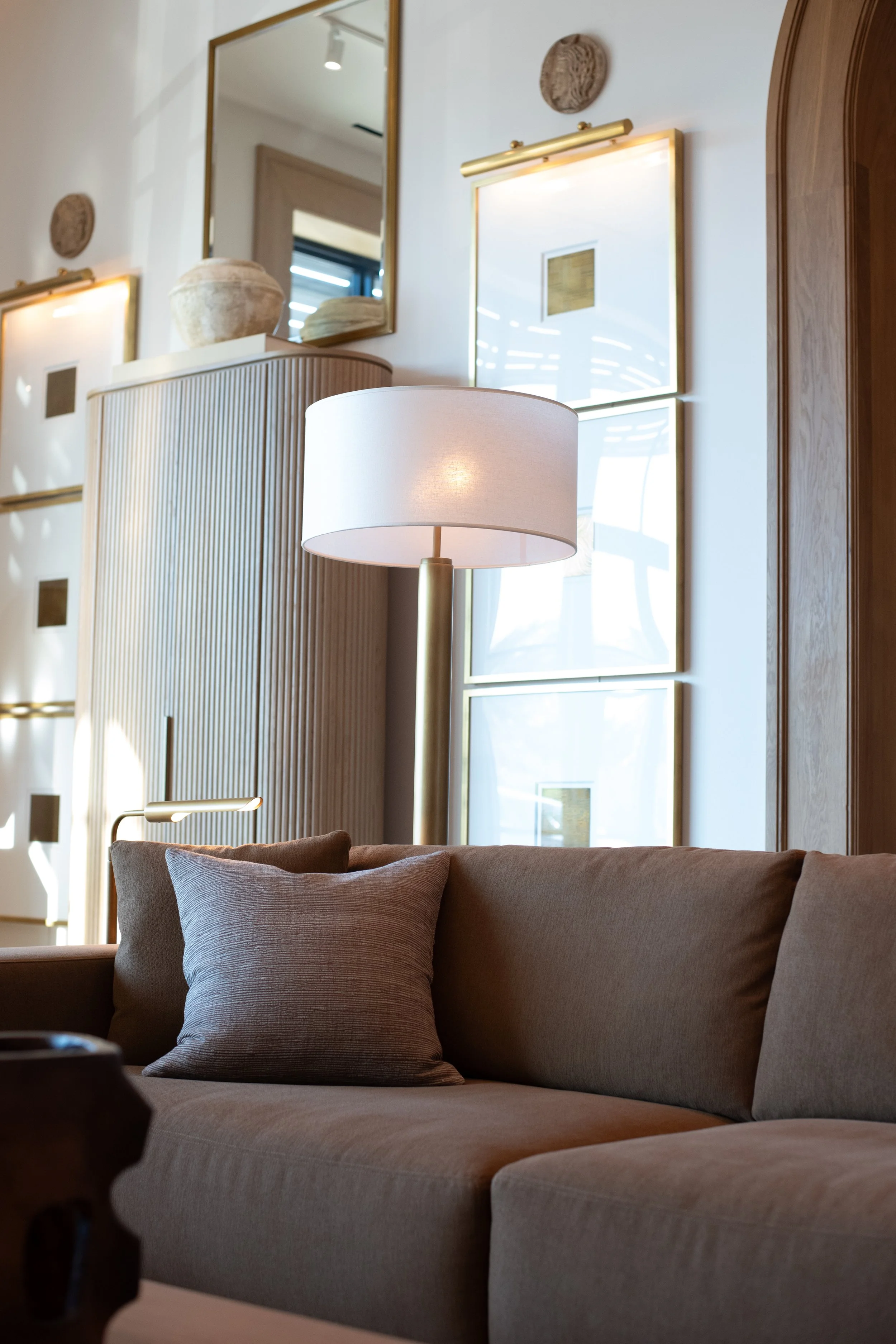 Living room with a beige sofa and cushions, a white lampshade, wooden walls, decorative wall art, a large mirror, and sunlight coming through windows.