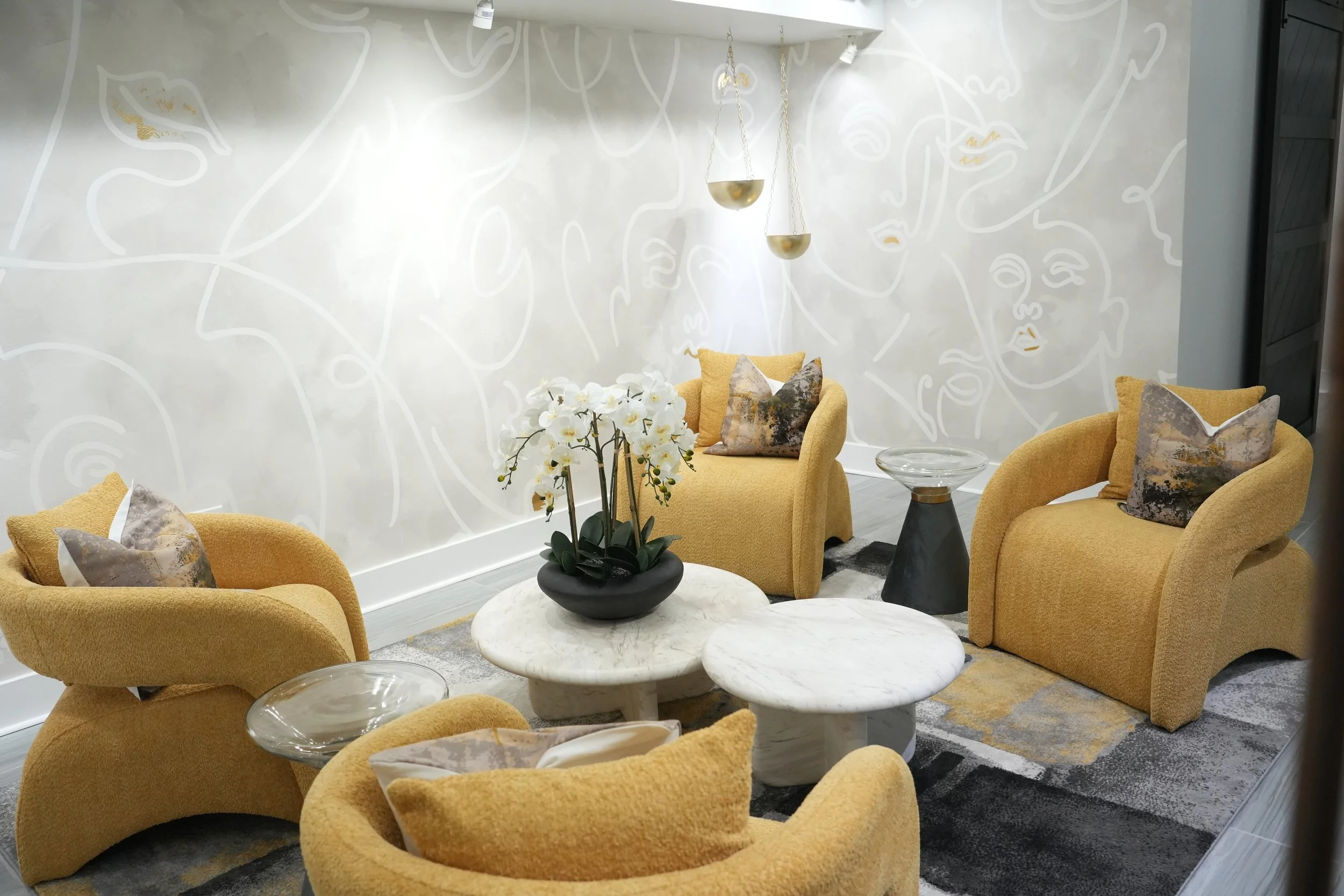 A modern lounge area with yellow upholstered armchairs and matching pillows, two white marble-topped coffee tables, and a centerpiece of white orchids. The background features a wall with abstract line art faces and hanging gold and white pendant lights.