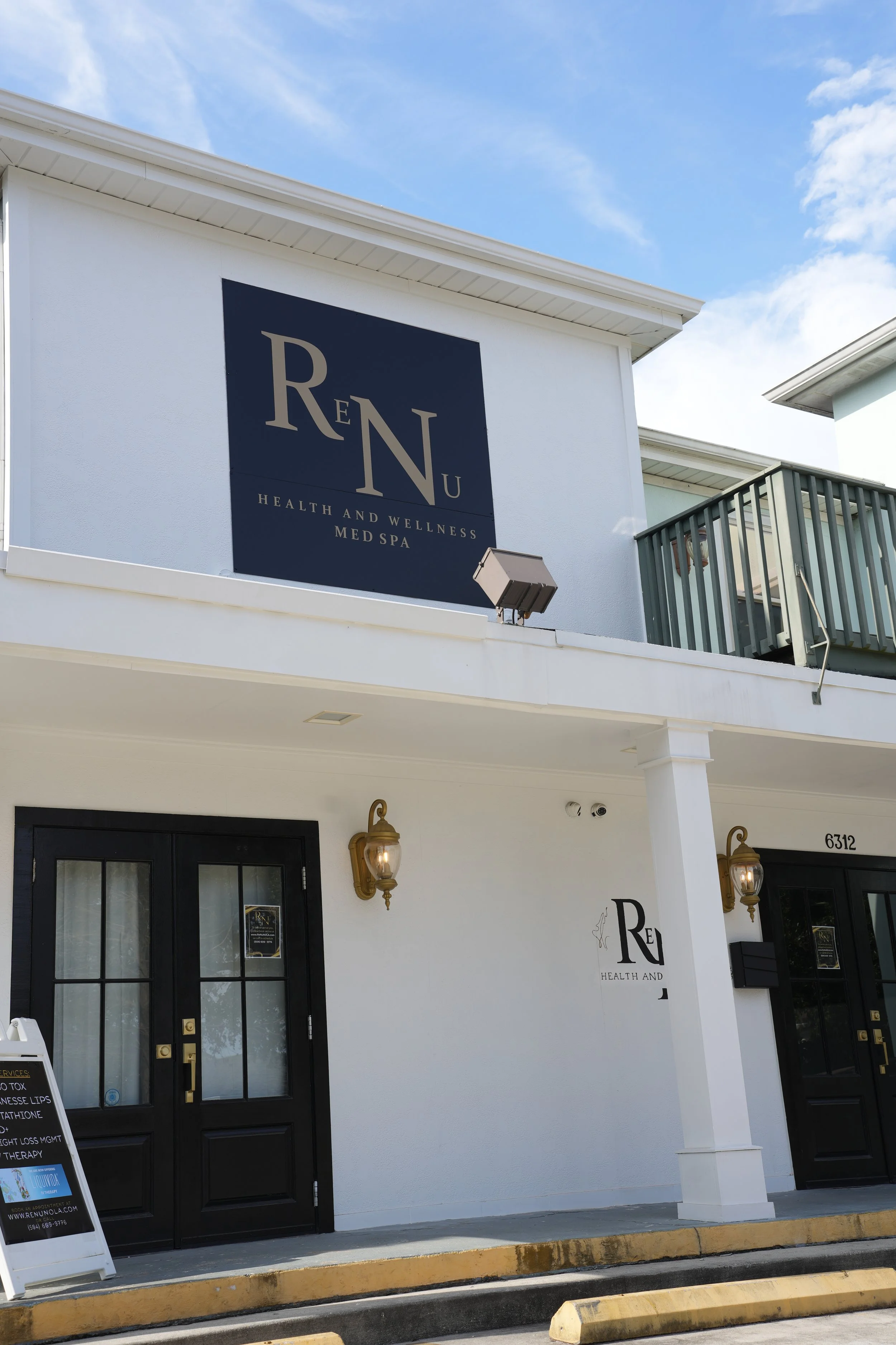 Exterior view of a health and wellness med spa named 'ReNu' with black double doors, outdoor lamps, and signage on a white building.