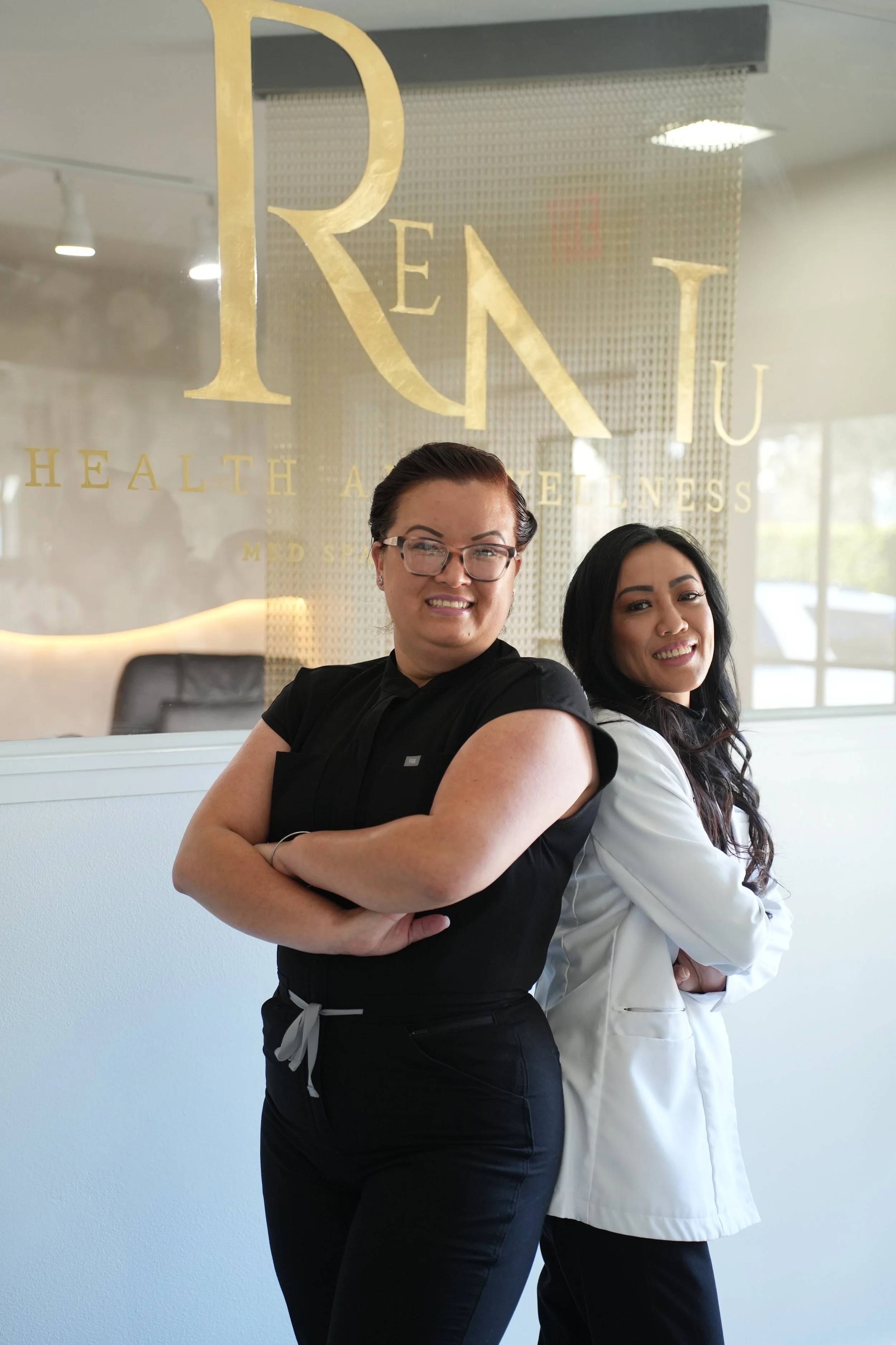 Two women standing back to back inside a wellness center, smiling at the camera. Behind them is a glass wall with gold lettering that reads 'Pen Itu' and 'Health and Wellness'.