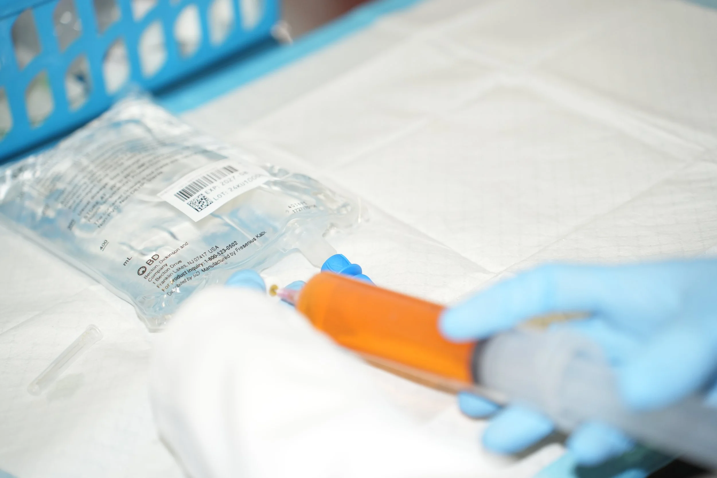Person wearing blue gloves holding a syringe filled with orange liquid near a bag of medical supplies on a white surface