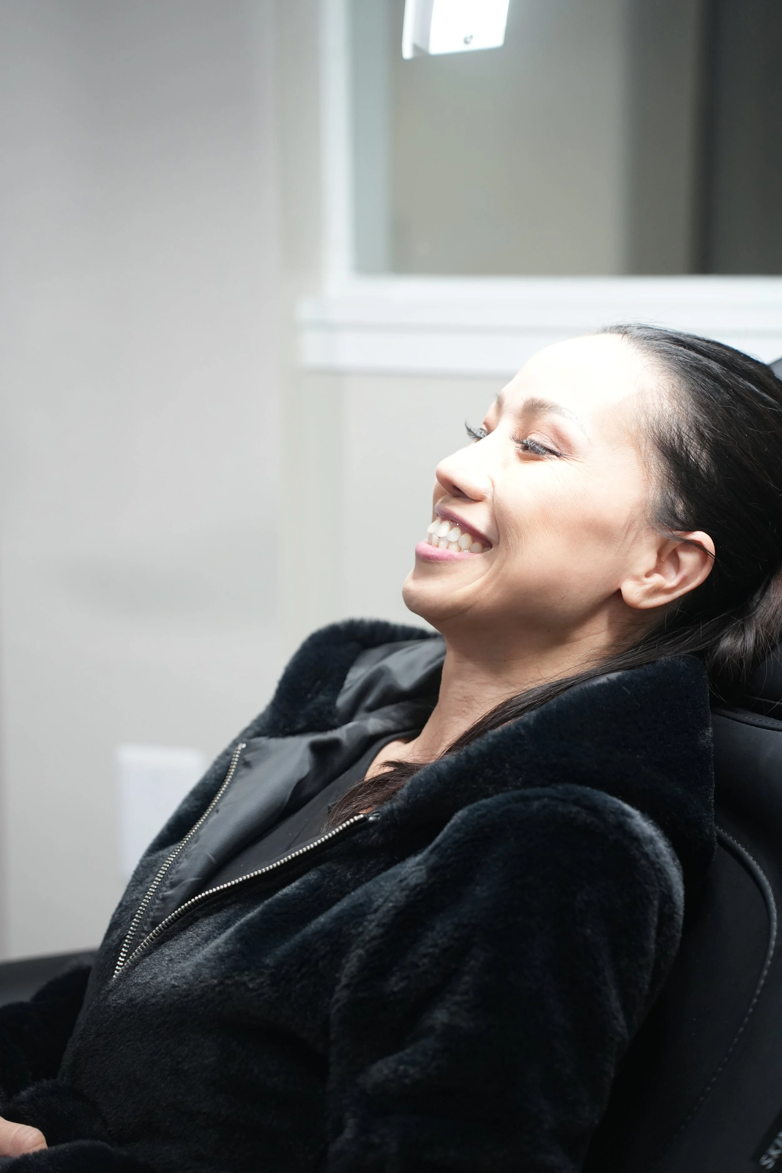 A woman with long dark hair smiling with her eyes closed, sitting in a chair in front of a light-colored wall with a window in the background.