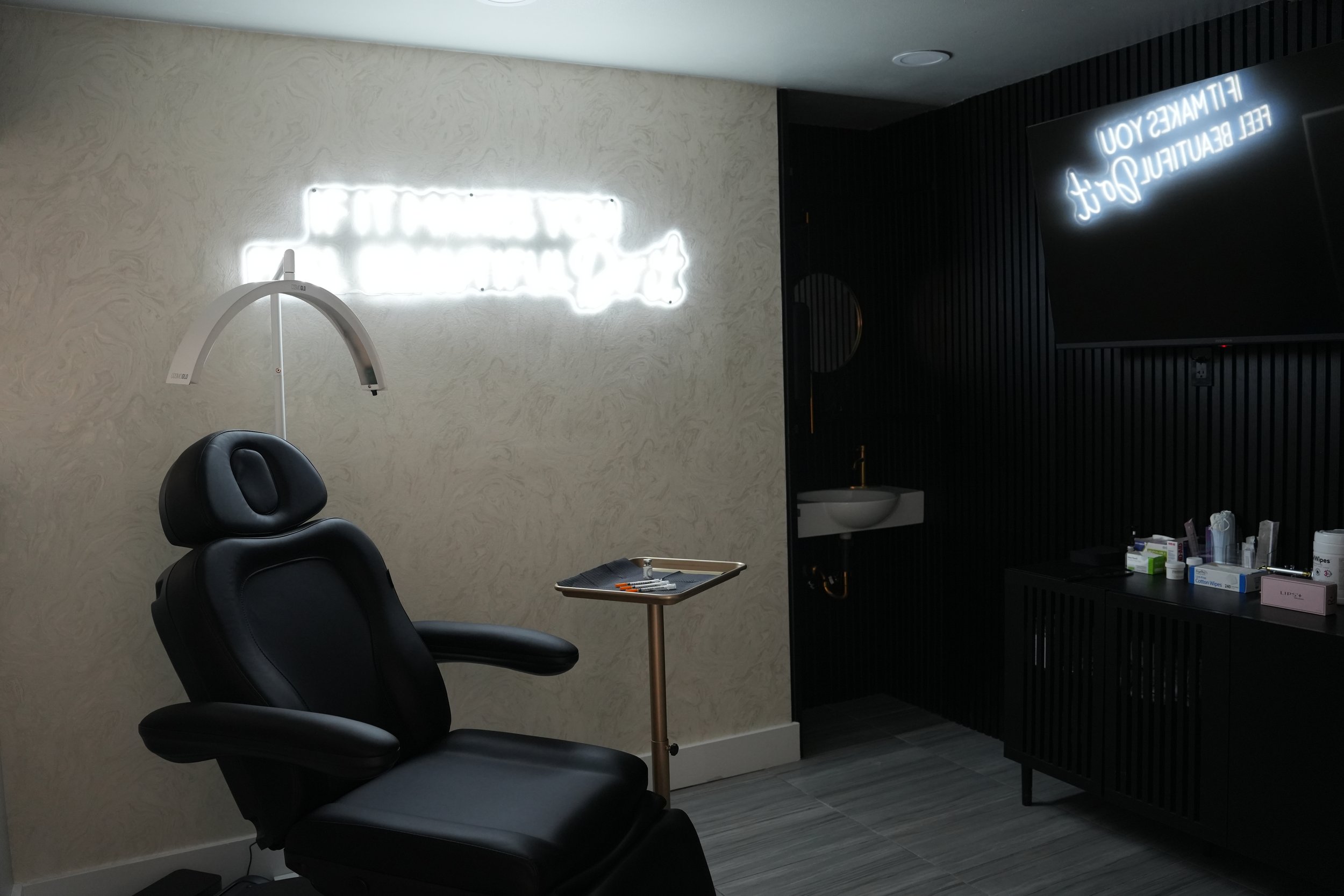 A dentist's office with a black dental chair, a small tray with dental tools, a black cabinet with medical supplies, and neon signs on the wall that read 'FILL MAKES YOU FEEL BEAUTIFUL' and other words.