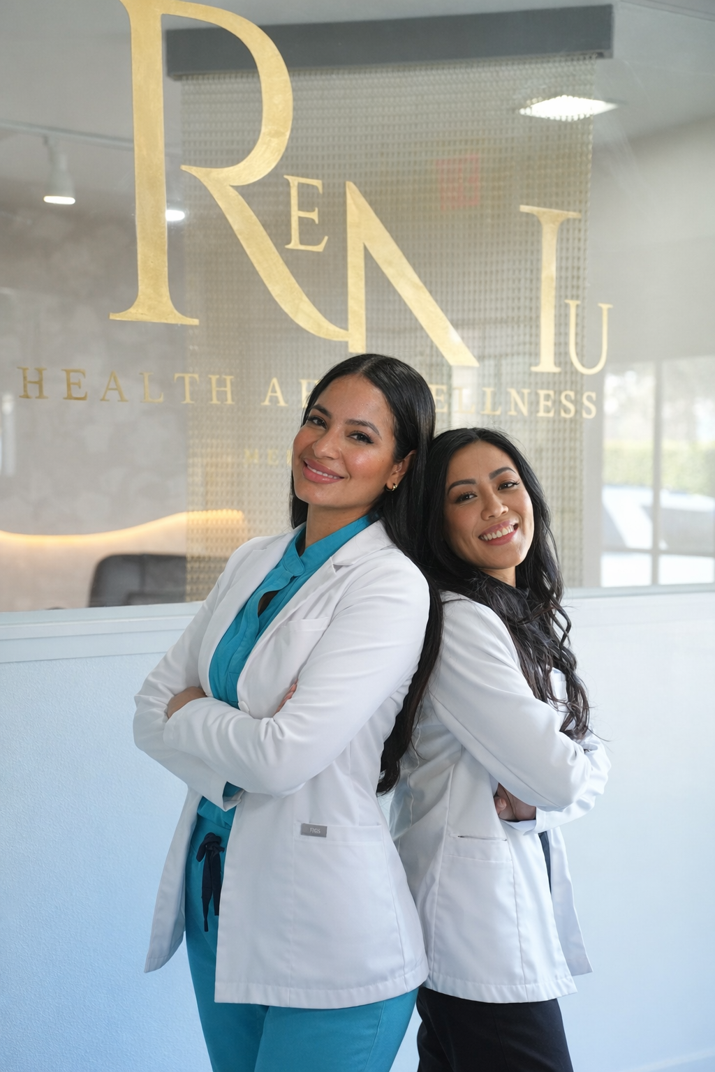 Two women in medical scrubs and white lab coats standing back-to-back, smiling with arms crossed, inside a clinic with a glass wall displaying a sign that reads "REN IU HEALTH AND WELLNESS."