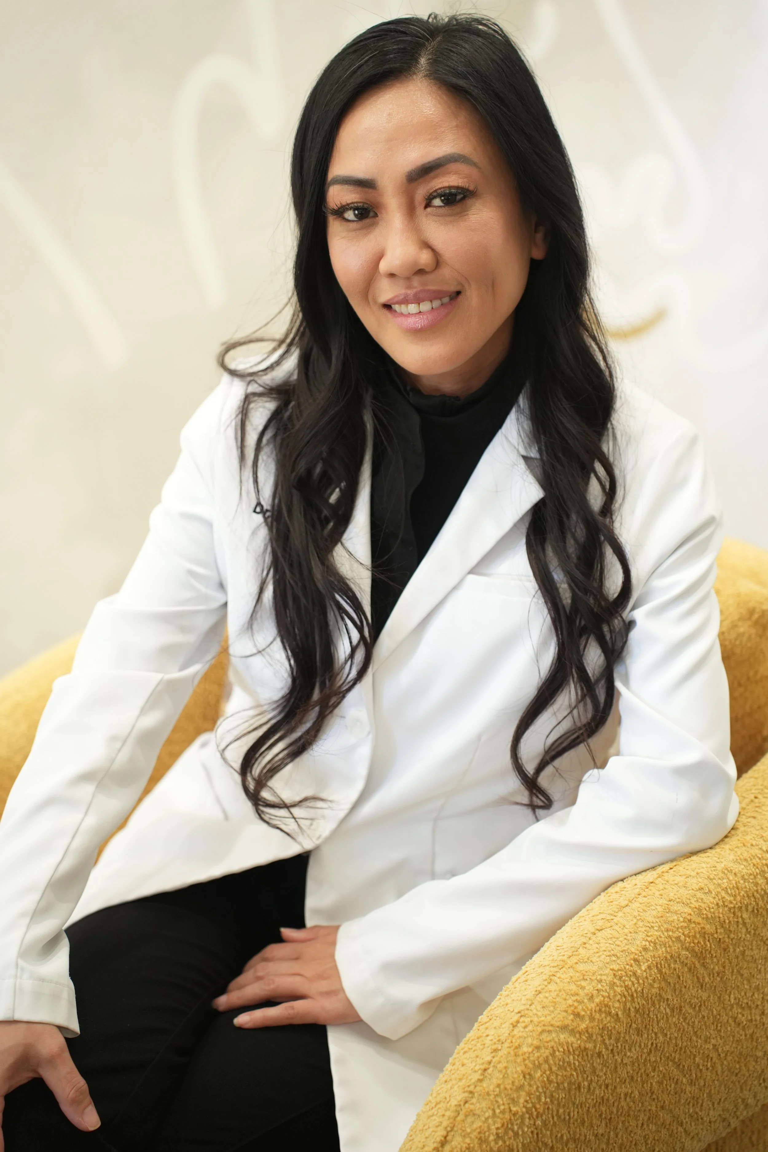 A woman with long dark hair, wearing a white coat, sitting on a yellow chair, smiling at the camera.