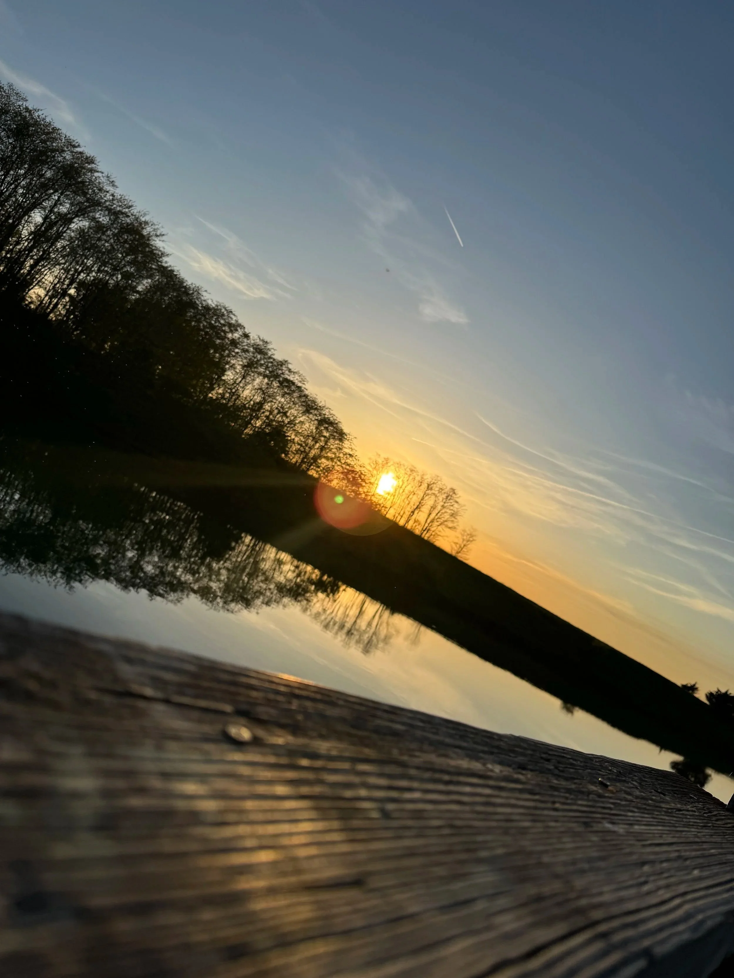 Sunset over a pond with reflections of trees, blue sky with clouds and contrail, viewed from a wooden surface in the foreground.
