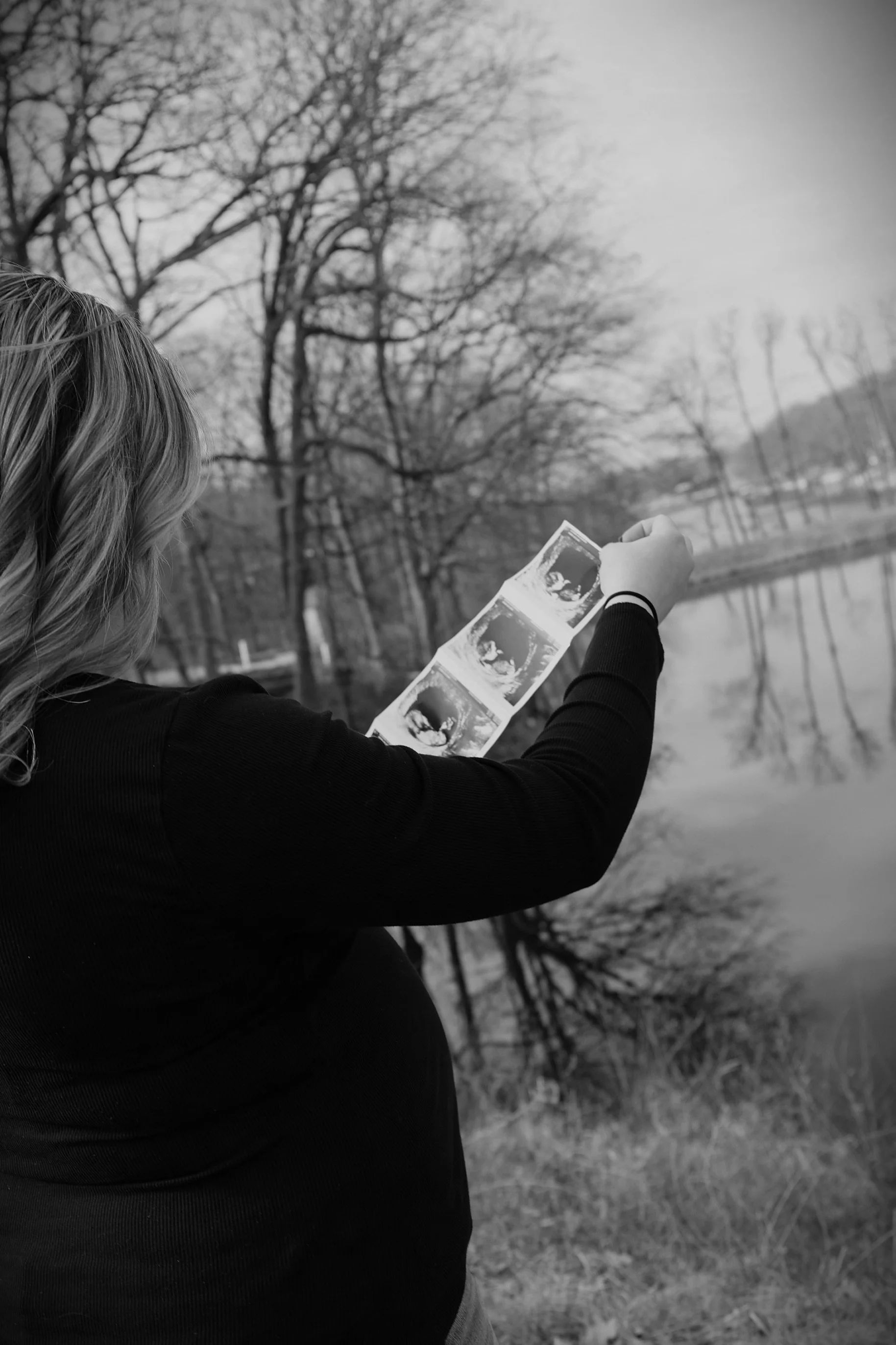 A woman with blonde hair holding a strip of four ultrasound photos near a body of water, with leafless trees in the background.