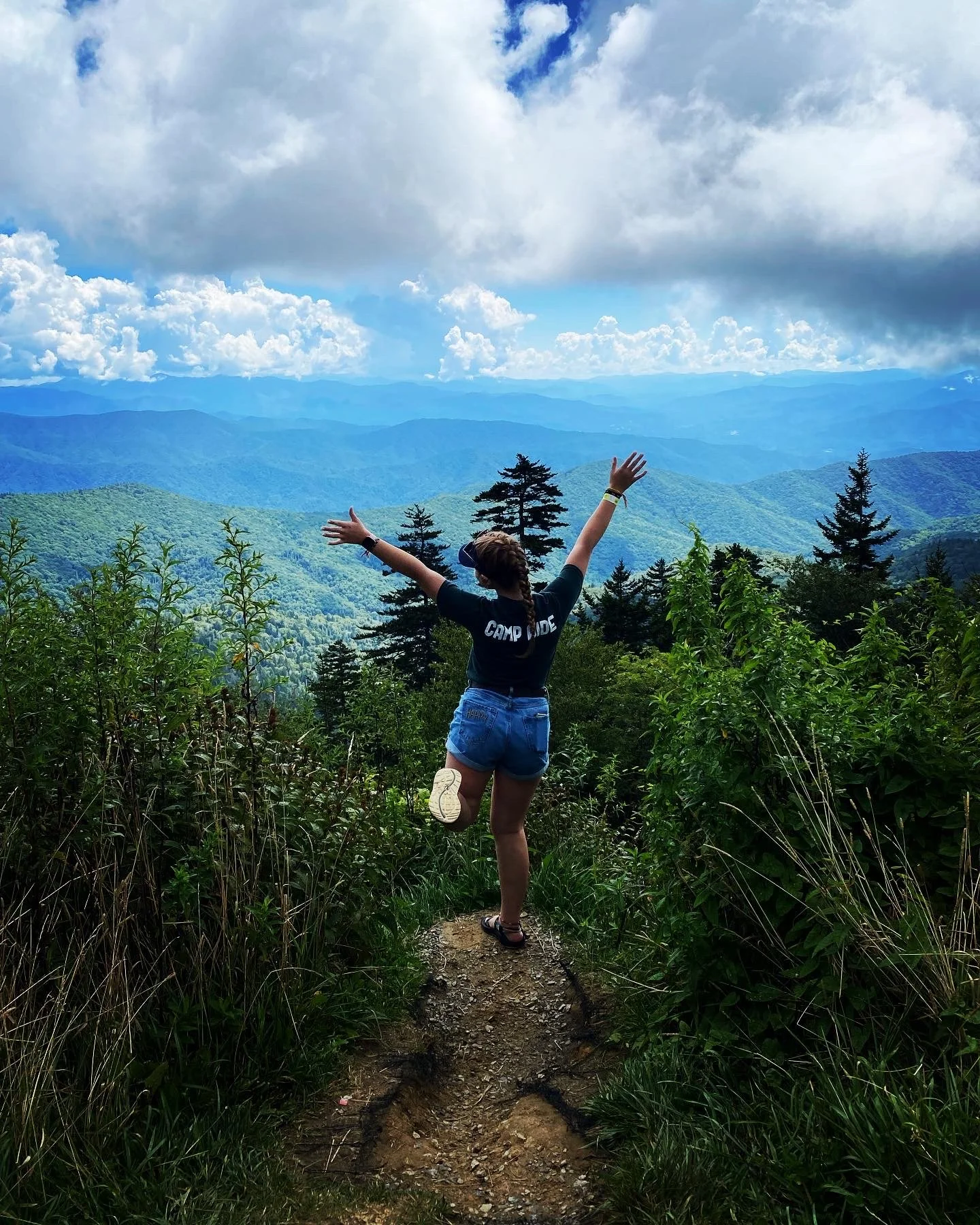 A person with braided hair wearing a black T-shirt and denim shorts standing on a trail with arms raised, overlooking a mountain range with trees and clouds in the sky.