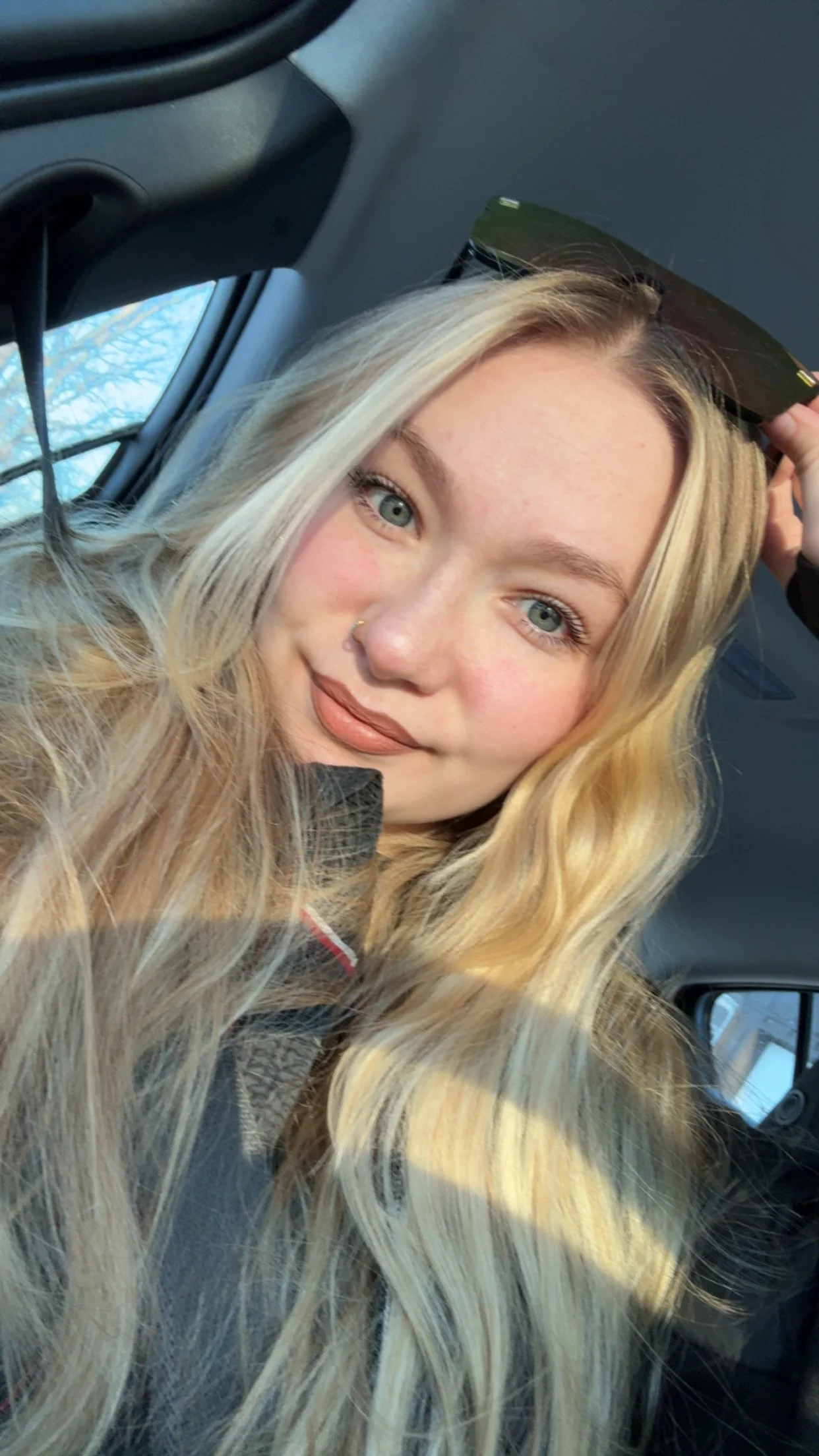 A young woman with long wavy blonde hair and blue eyes sitting inside a vehicle, holding sunglasses on her forehead.