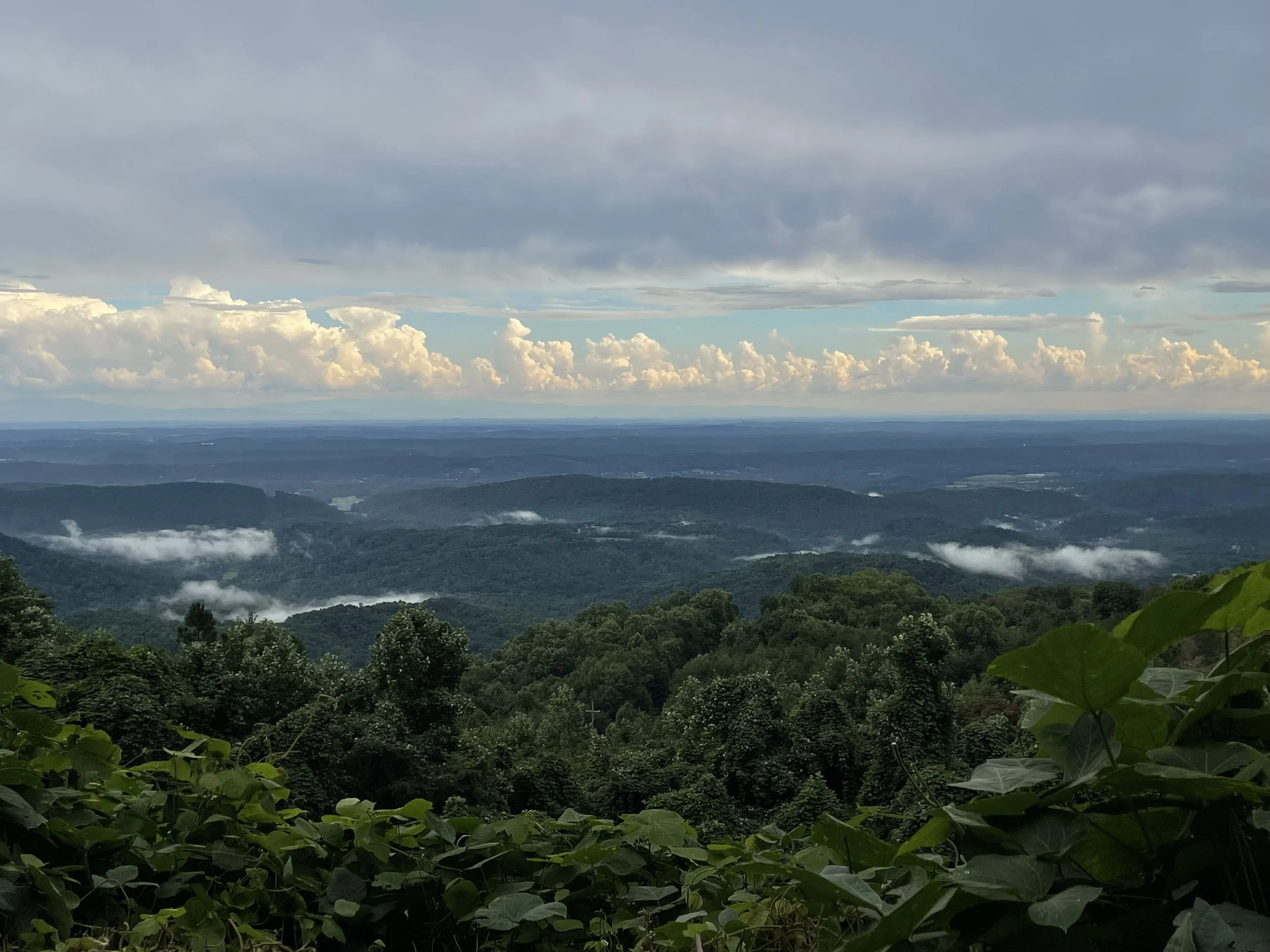 Scenic view of green, forested mountains with clouds floating between the hills under a partly cloudy sky.