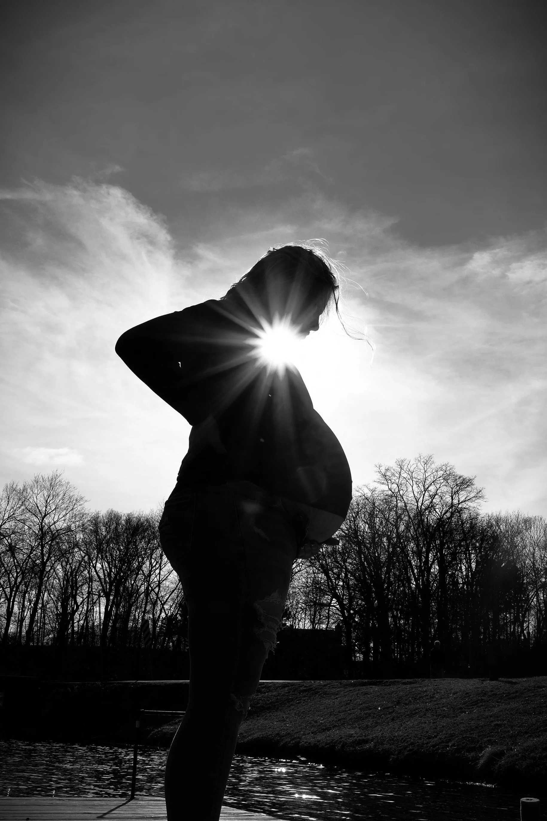 Silhouette of a pregnant woman standing outdoors near water with trees in the background, backlit by the sun, creating a sunburst effect.