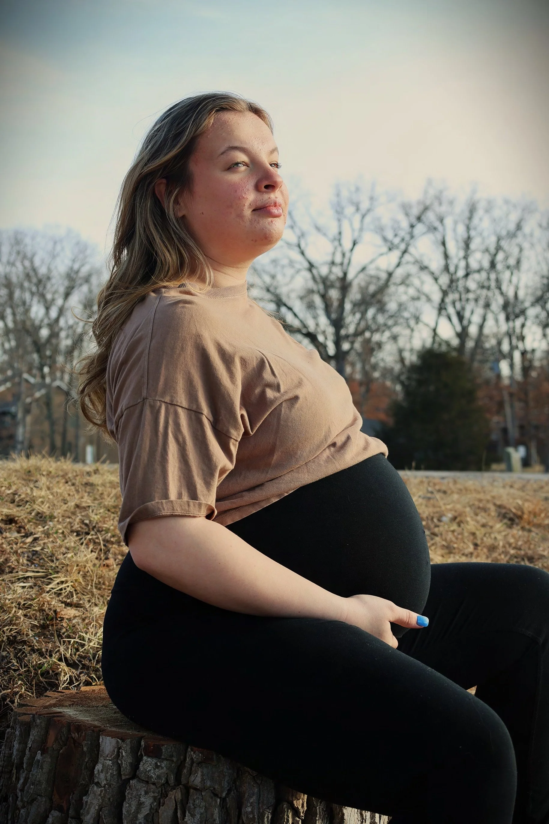 A pregnant woman sitting outdoors on a tree stump, looking into the distance with a serene expression, trees in the background with no leaves, and a soft, natural light.