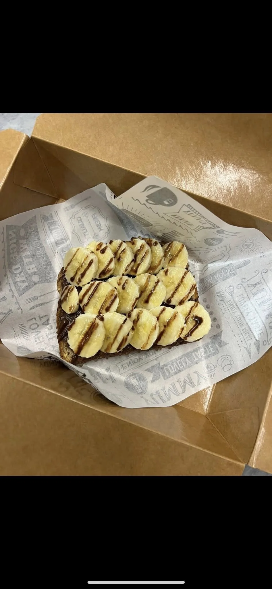 A banana and chocolate toast with banana slices and chocolate syrup in a brown cardboard box