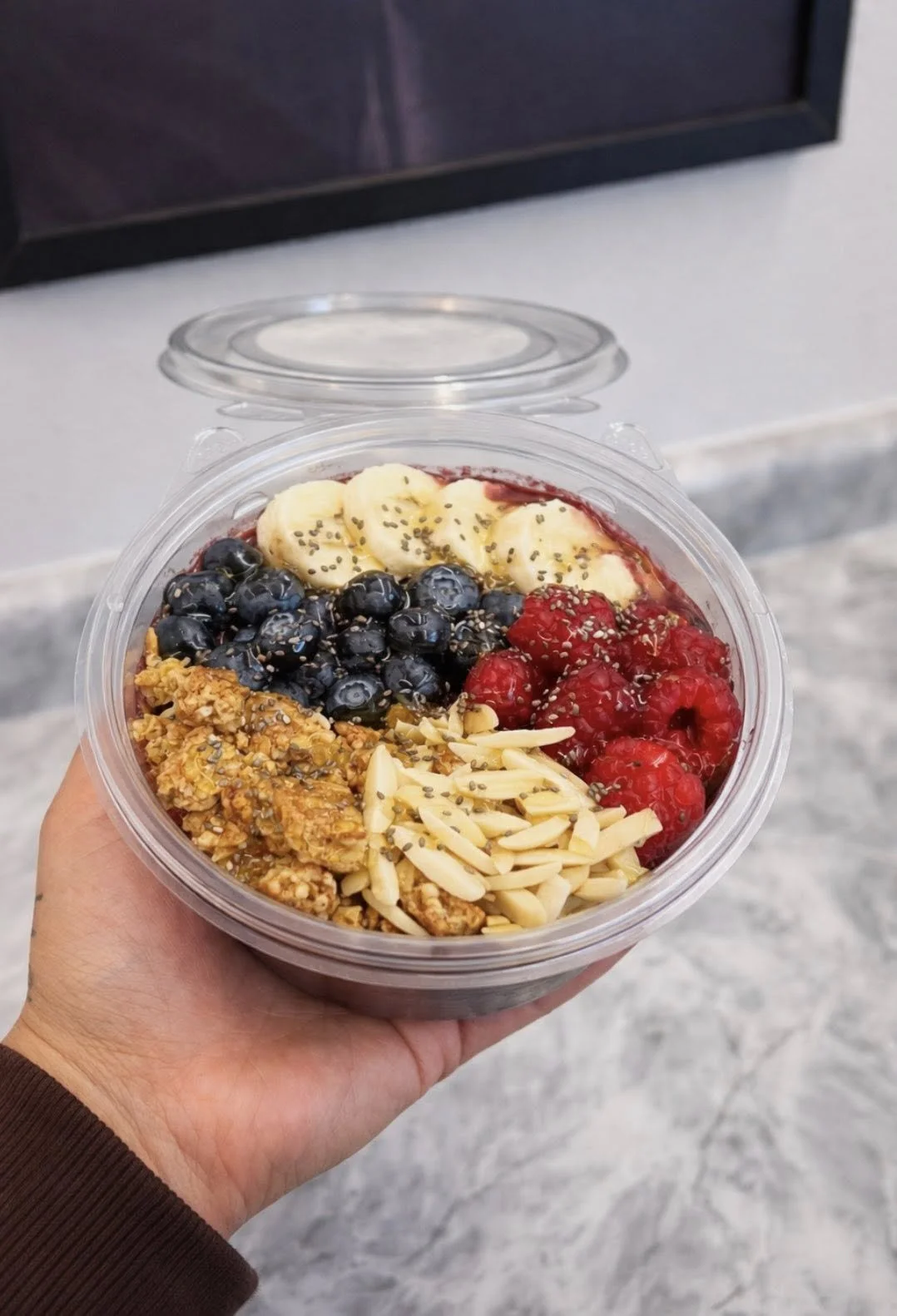 A hand holding a clear plastic container with a colorful layered smoothie topped with banana slices, blueberries, raspberries, granola, slivered almonds, and chia seeds. The background appears to be a gray wall and marble counter.