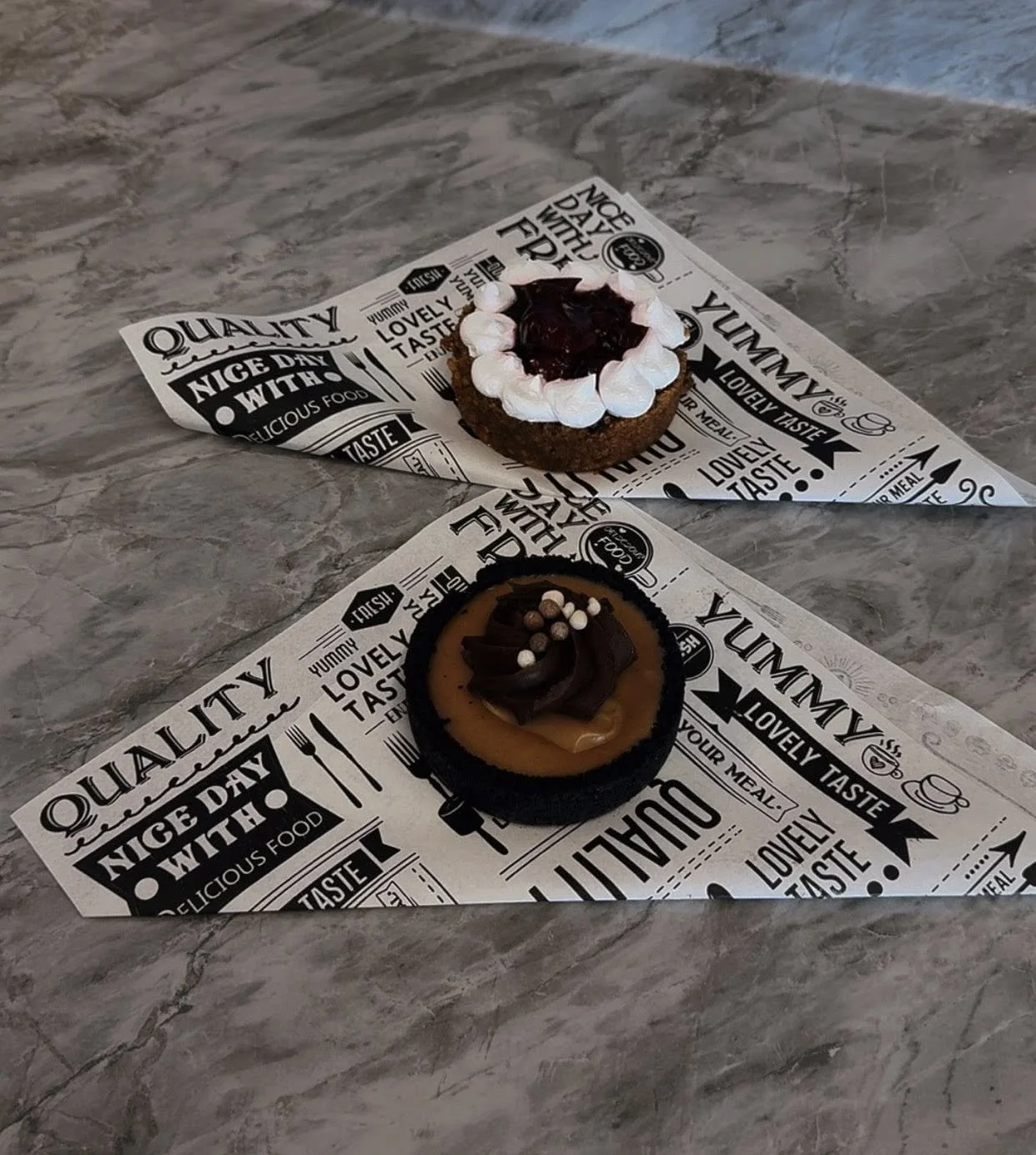 Two decorated cupcakes on black and white printed paper sheets placed on a marble countertop.