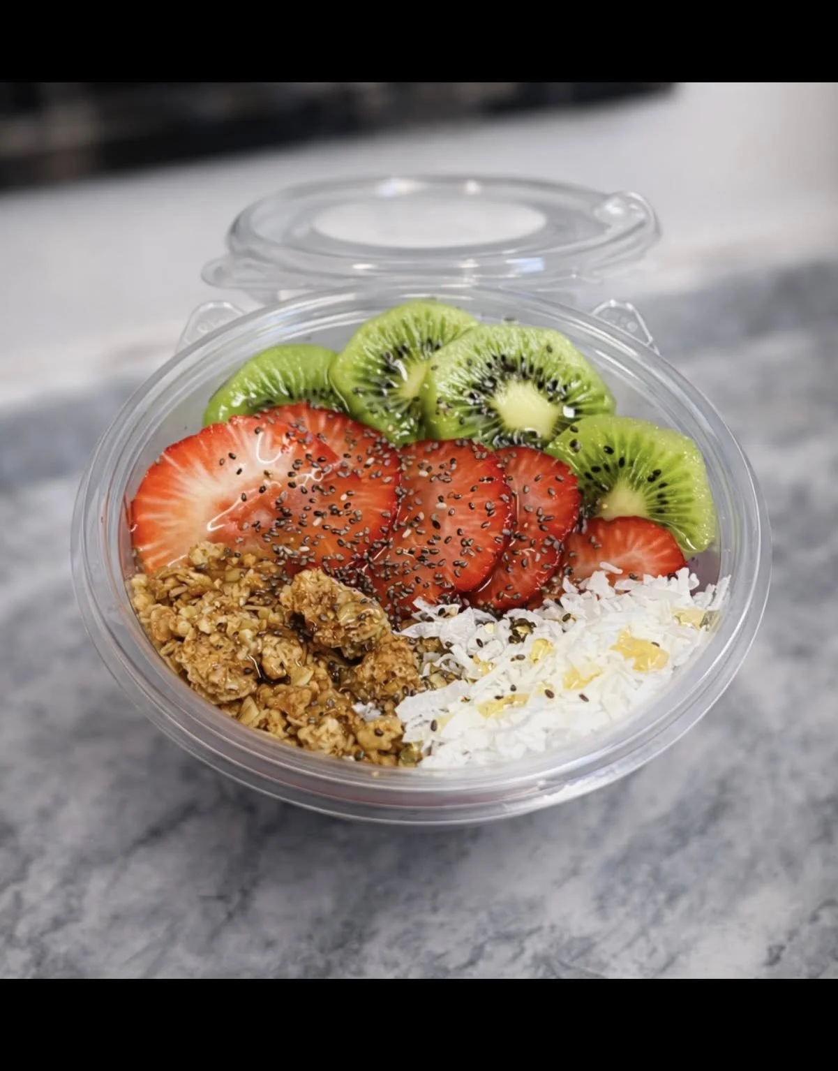 A clear plastic bowl with sliced strawberries, kiwi, granola, shredded coconut, and black sesame seeds on top.