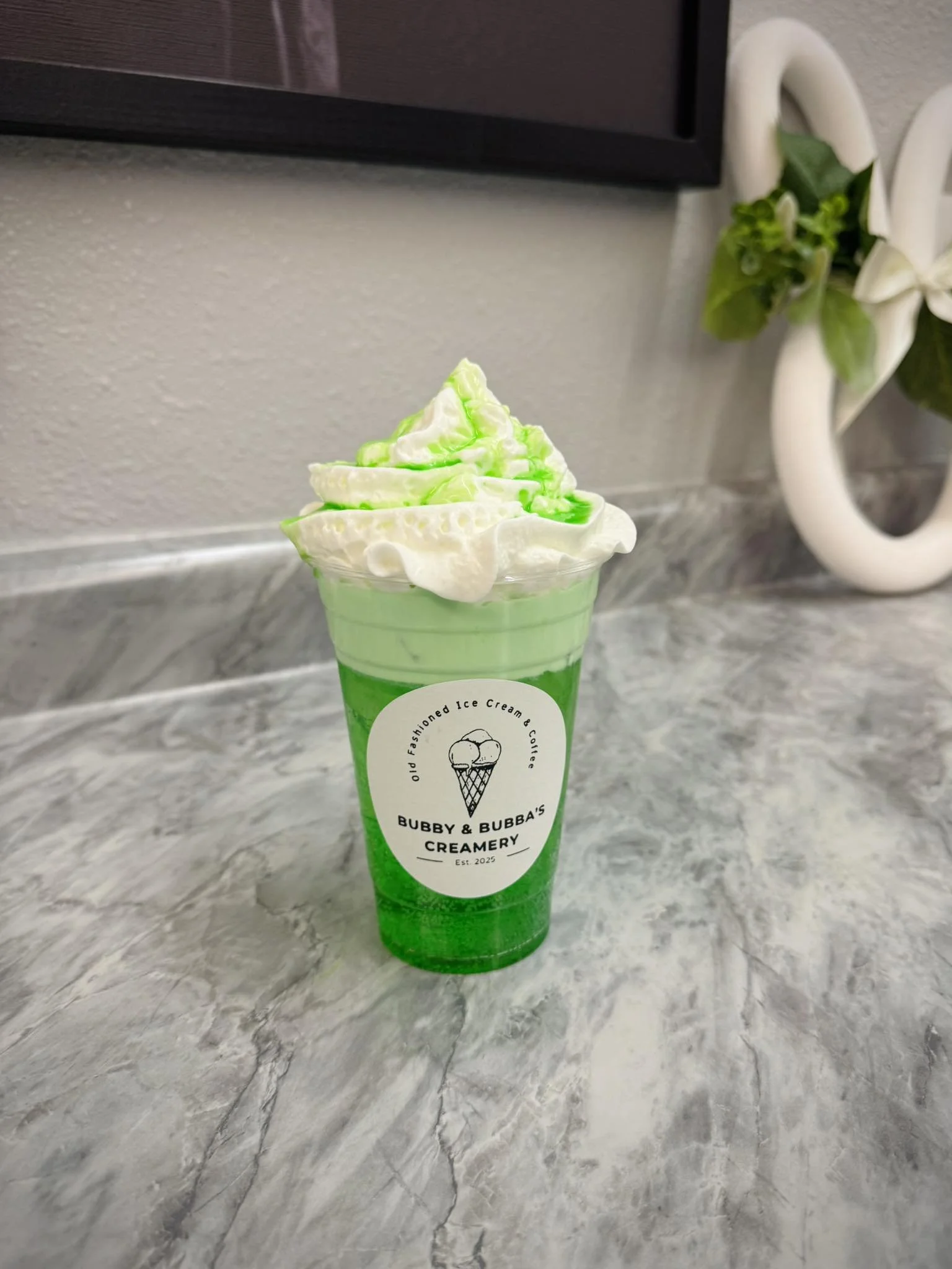 A green and white ice cream float in a clear cup with whipped cream on top, labeled Bubby & Bubba's Creamery, on a marble countertop.