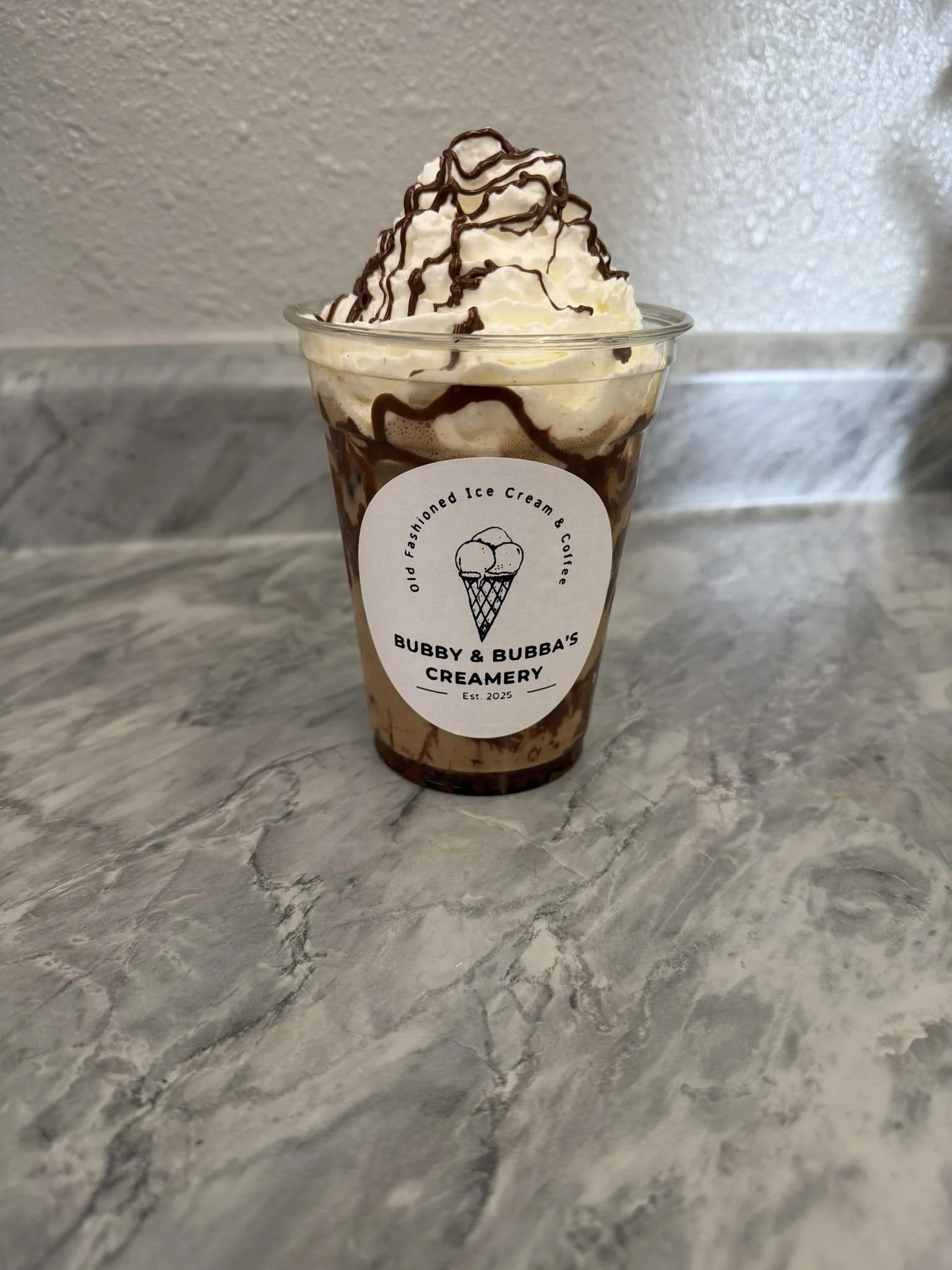 A clear plastic cup filled with ice cream topped with whipped cream and drizzled with chocolate syrup. The cup has a label with an ice cream cone logo and the text 'Bubby & Bubba's Creamery'.