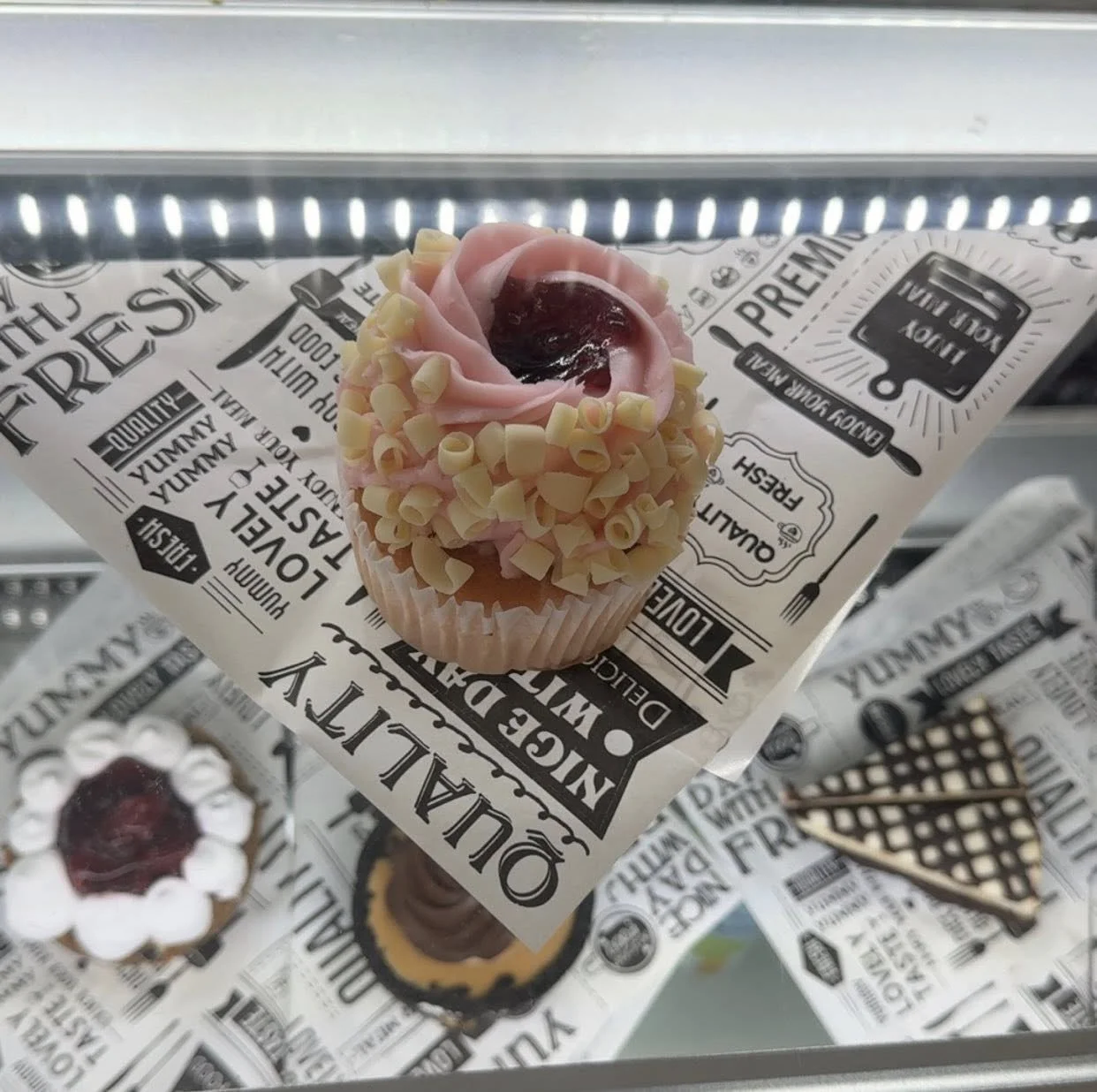 A cupcake decorated with pink frosting, white chocolate curls, and a cherry on top, displayed on a black and white paper wrapper inside a glass display case.