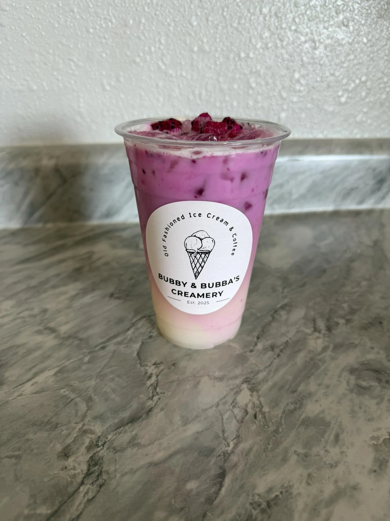 A cup of pink and white ice cream with berry topping from Bubby & Bubba's Creamery, placed on a gray marble surface against a textured wall.