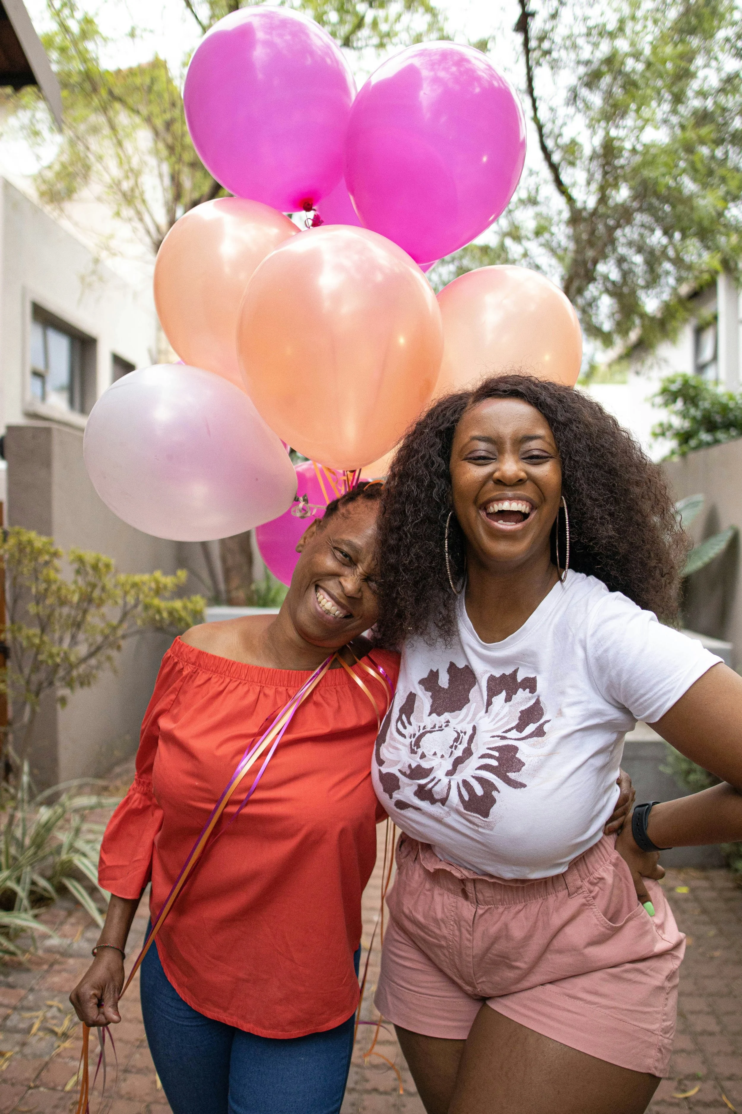 Two women smiling and laughing outdoors, holding pink, peach, and white balloons, surrounded by trees and buildings.
