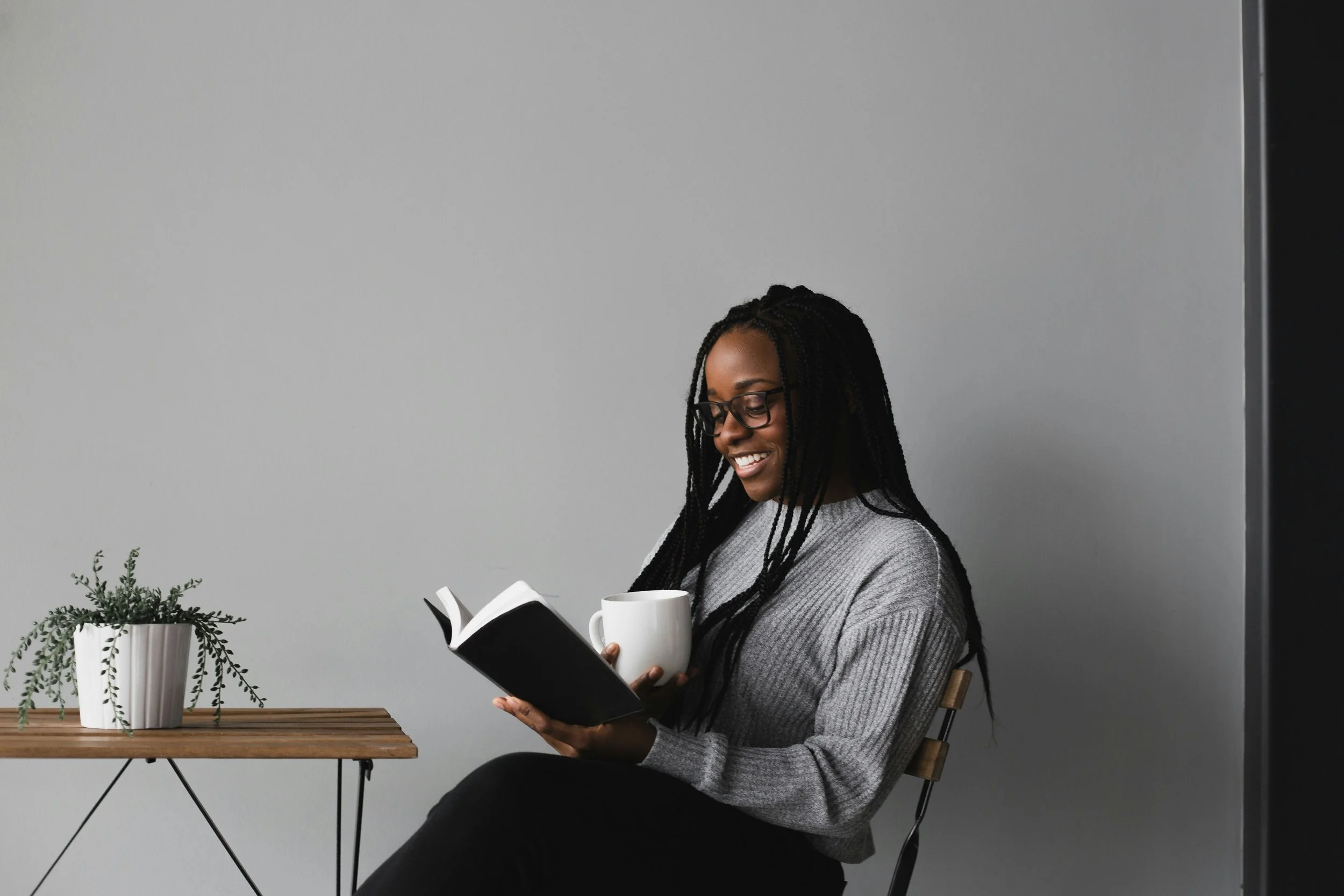 A woman with glasses and braided hair is sitting on a chair, smiling while reading a book and holding a mug. There is a small wooden table with a potted plant beside her, against a plain gray background.