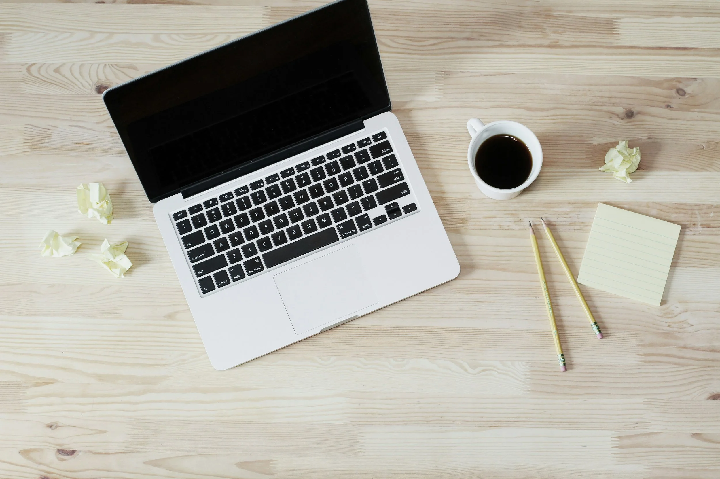 A desk with a silver laptop, a cup of black coffee, two yellow pencils, a yellow sticky note pad, and crumpled pieces of paper on a light wooden surface.