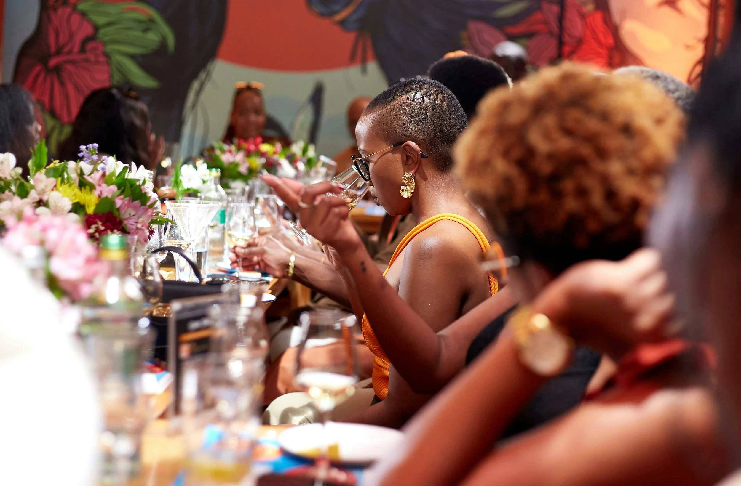 Women seated at a dinner table drinking wine, surrounded by floral centerpieces, at a social gathering or celebration.