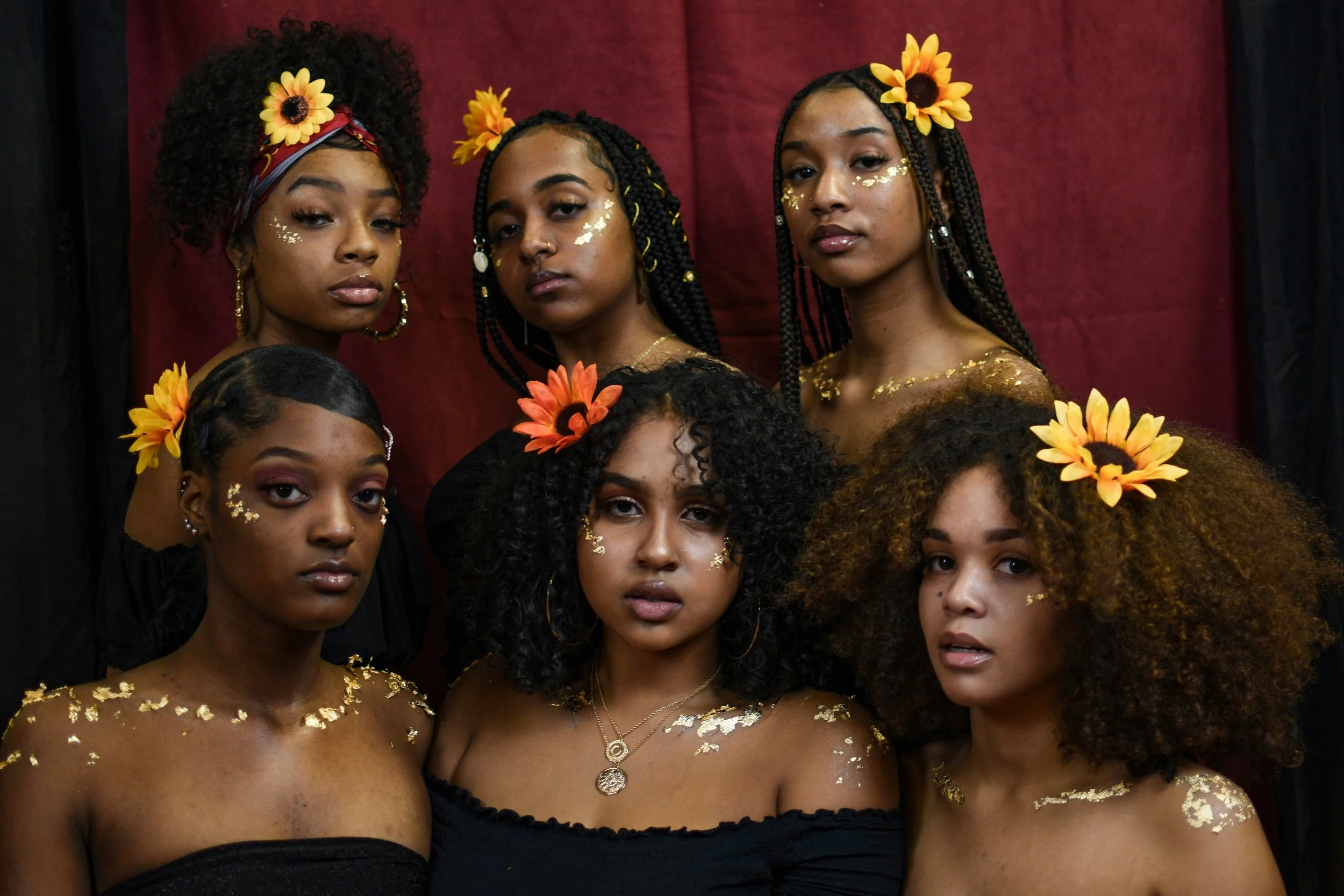 Six women with gold leaf makeup and sunflower accessories, posing against a maroon background.