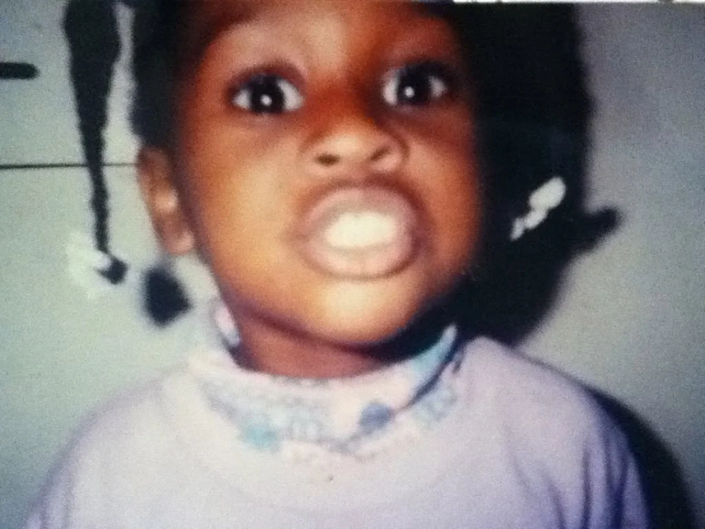 Close-up of a young girl making a fierce face with her teeth showing, wearing a pastel-colored shirt with a floral collar and two braids with white bows, against a neutral background.