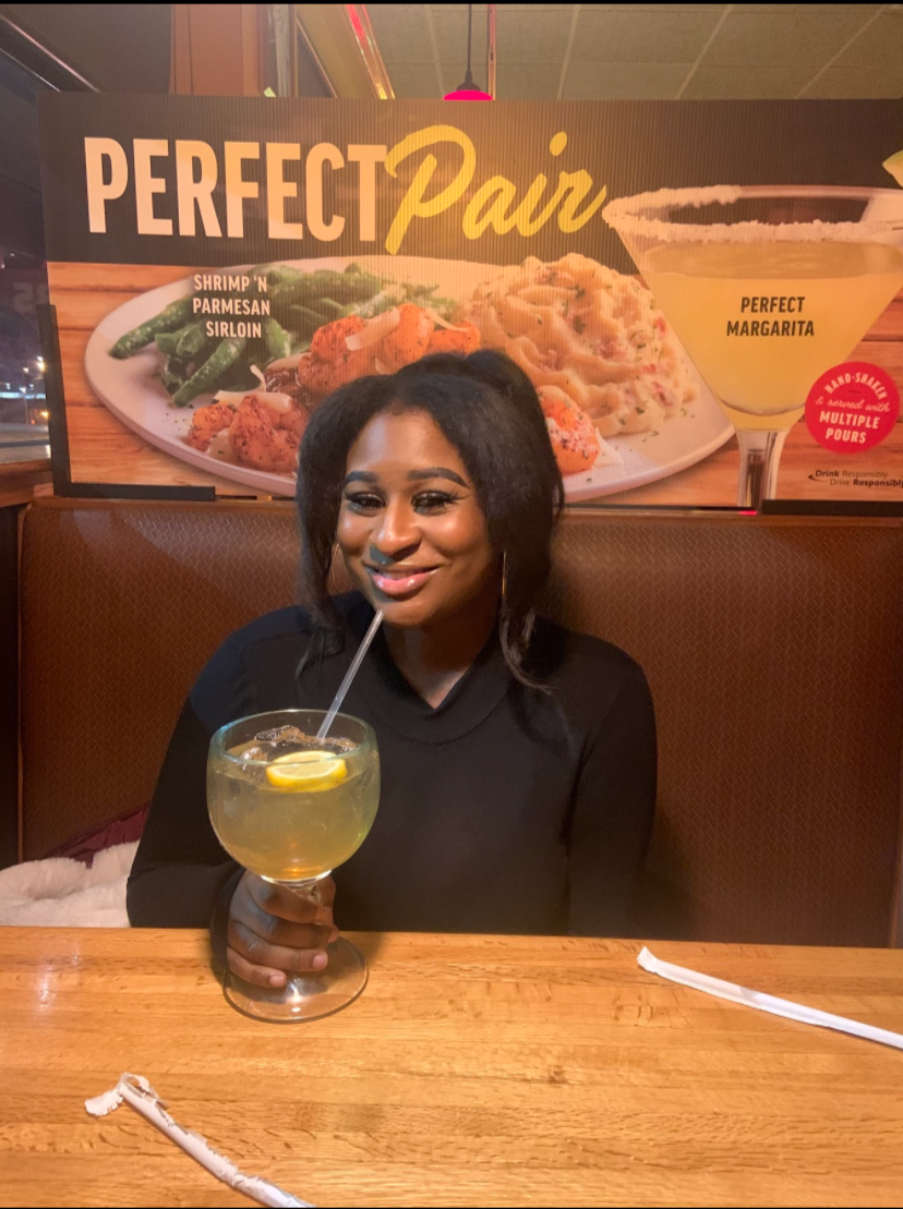 A woman with dark hair sitting at a wooden table, holding a large glass of lemonade with lemon slices, in a restaurant. Behind her is a sign that says "Perfect Pair" with images of shrimp 'n' parmesan sirloin, pasta, and a margarita.