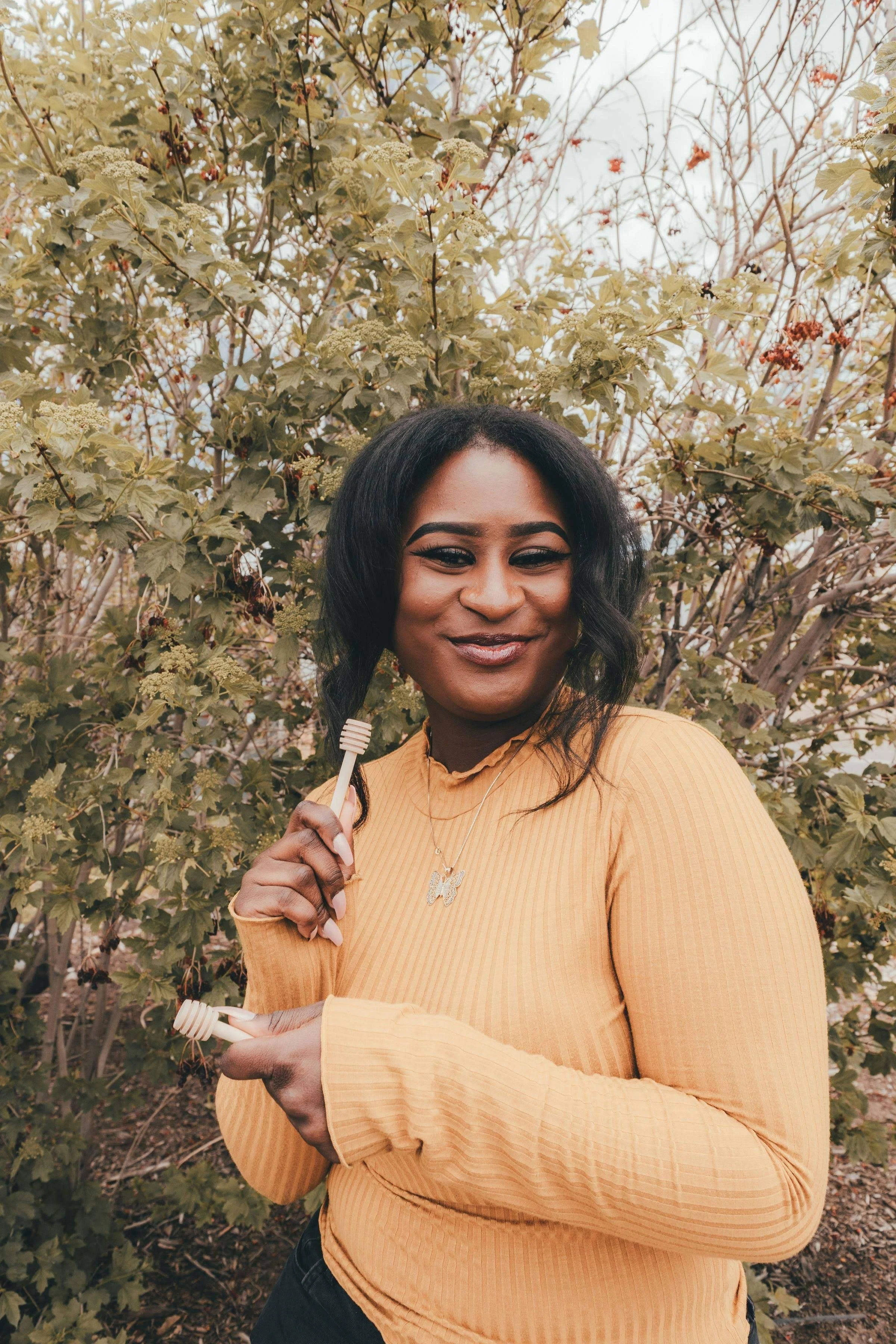 A woman with dark hair and a yellow long-sleeve shirt smiling outdoors, holding a wooden honey dipper, standing in front of green leafy bushes.