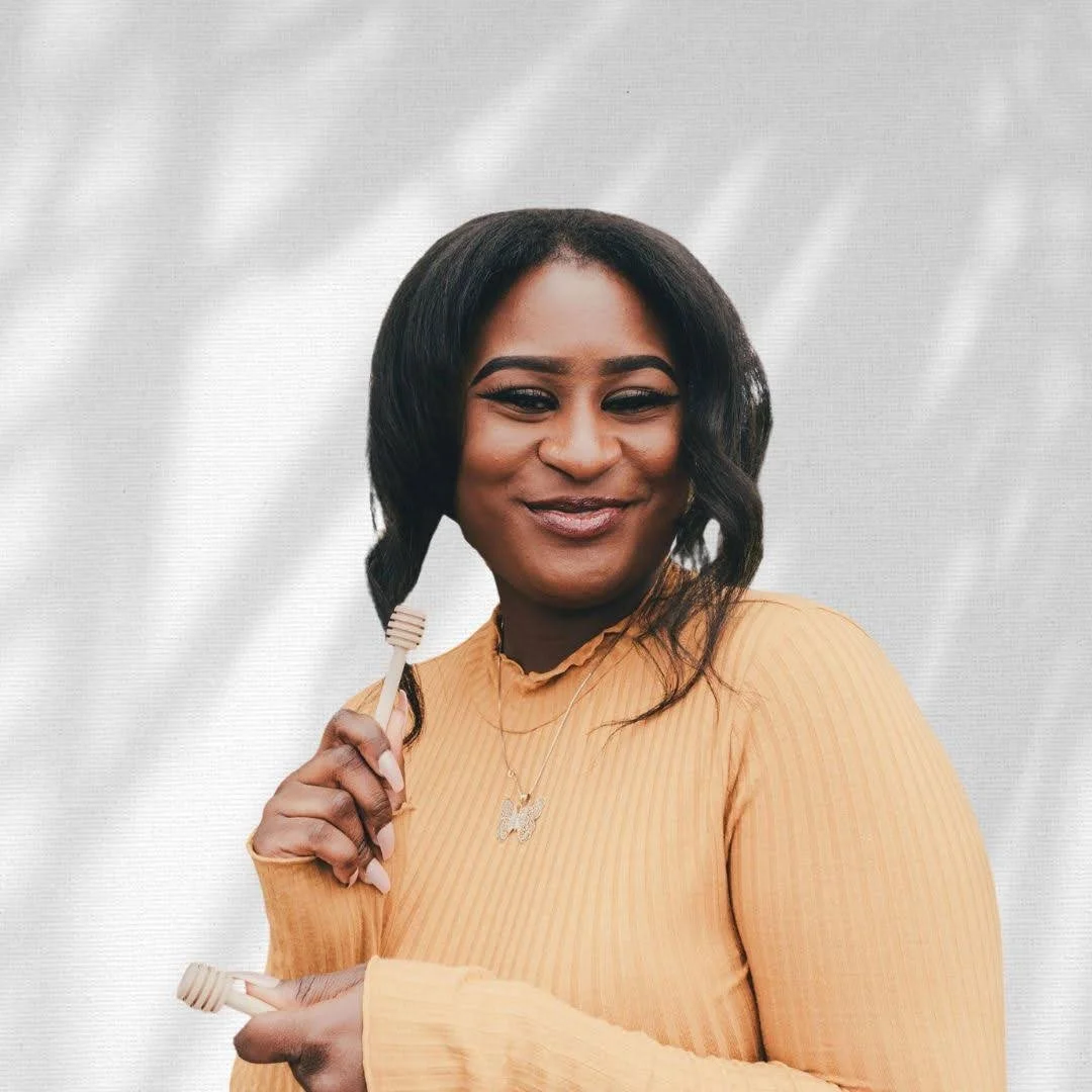 A smiling woman holding wooden honey dippers in her hands, wearing a peach-colored ribbed top, with a white and gray background.