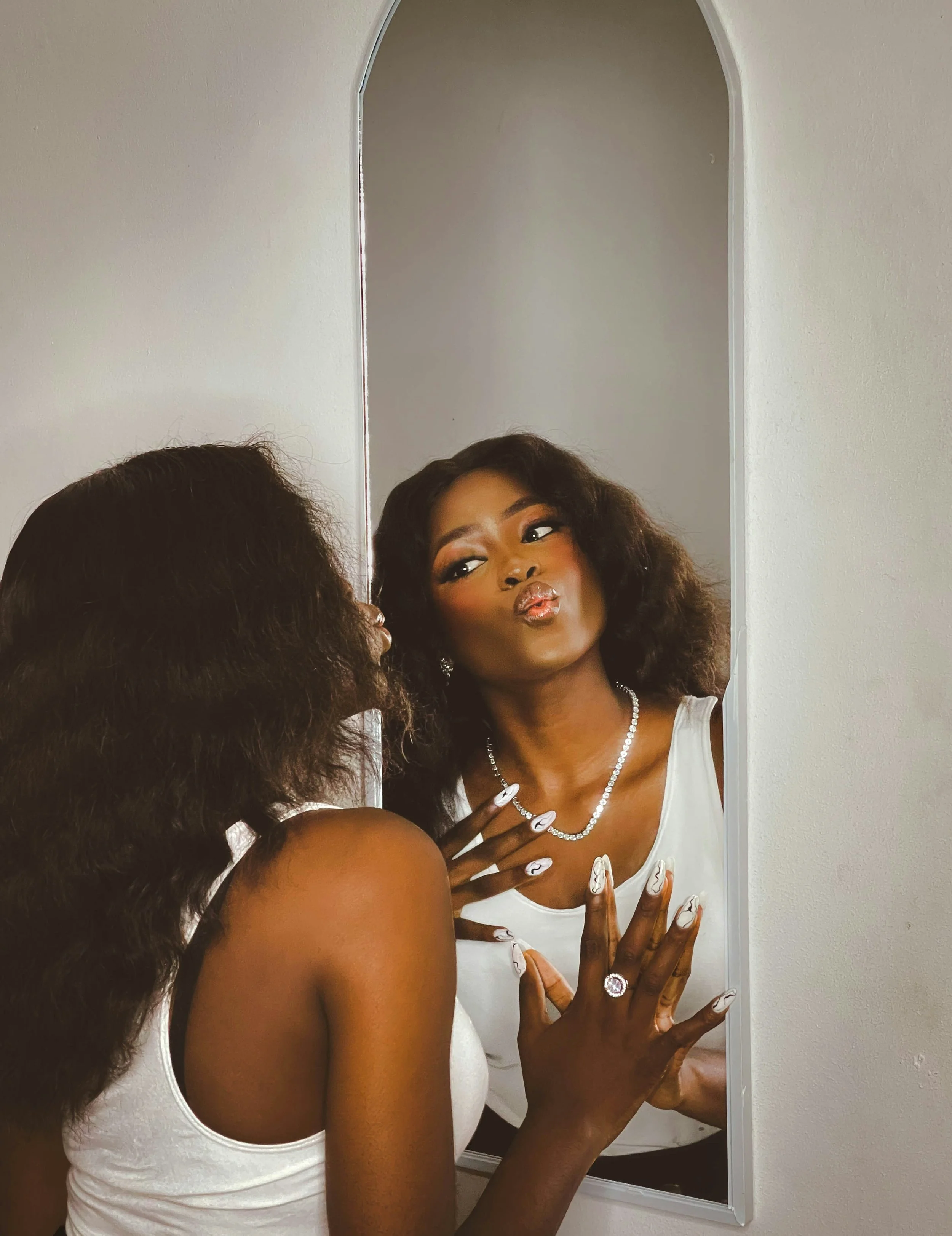 A woman with dark curly hair looking at herself in a mirror, pursing her lips, wearing a white top and jewelry.