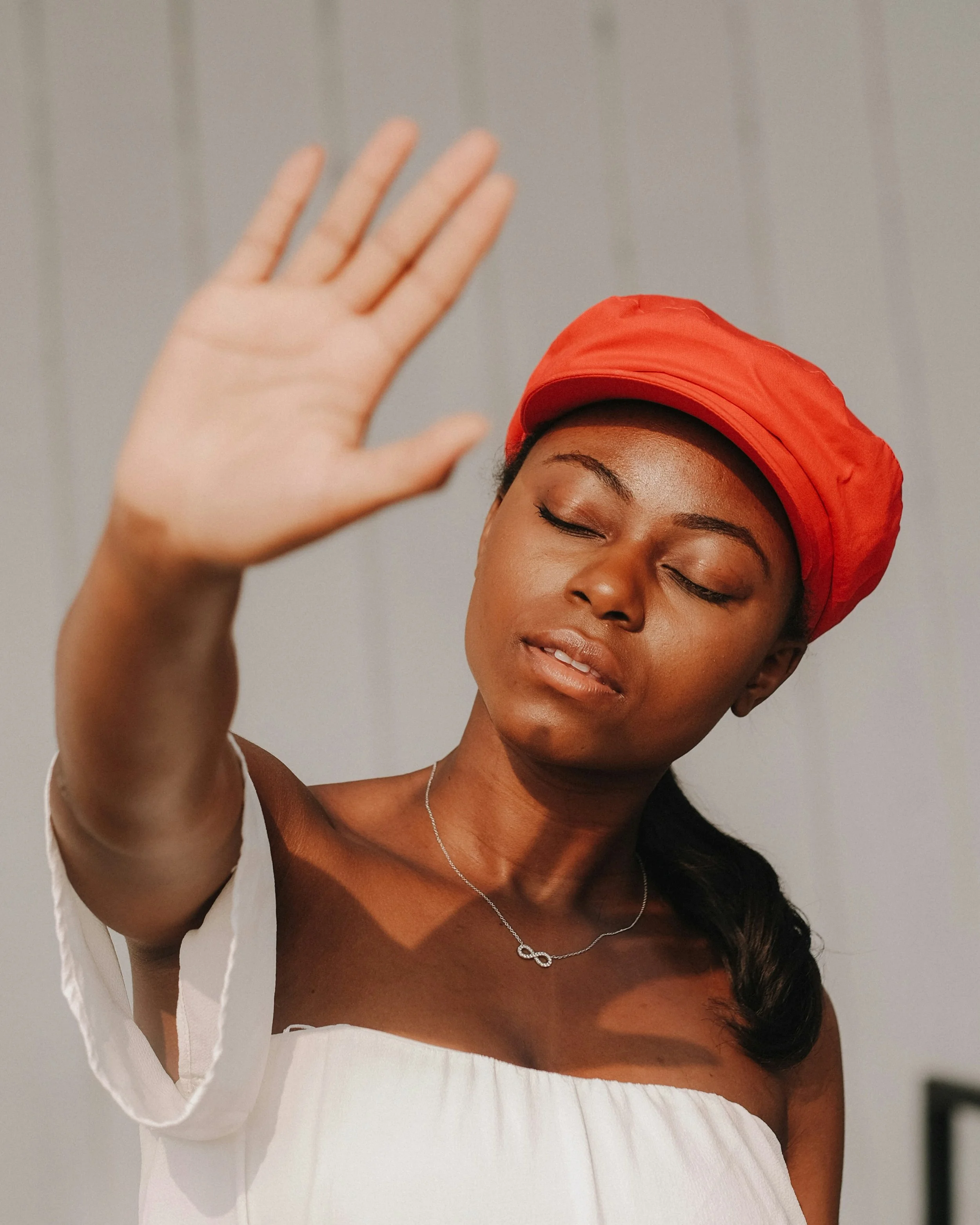 A young woman with dark hair in a ponytail, wearing a bright red headscarf and a white off-the-shoulder top, with eyes closed and hand raised in front of her face.