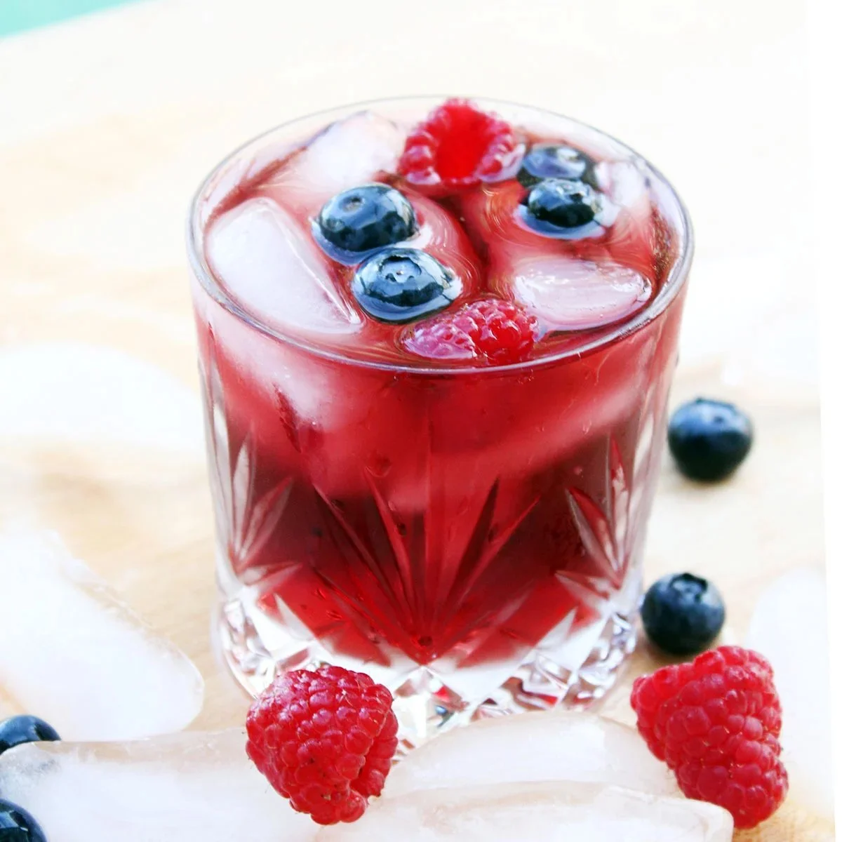 A glass of iced berry beverage with blueberries and raspberries on top, surrounded by additional berries and ice cubes.