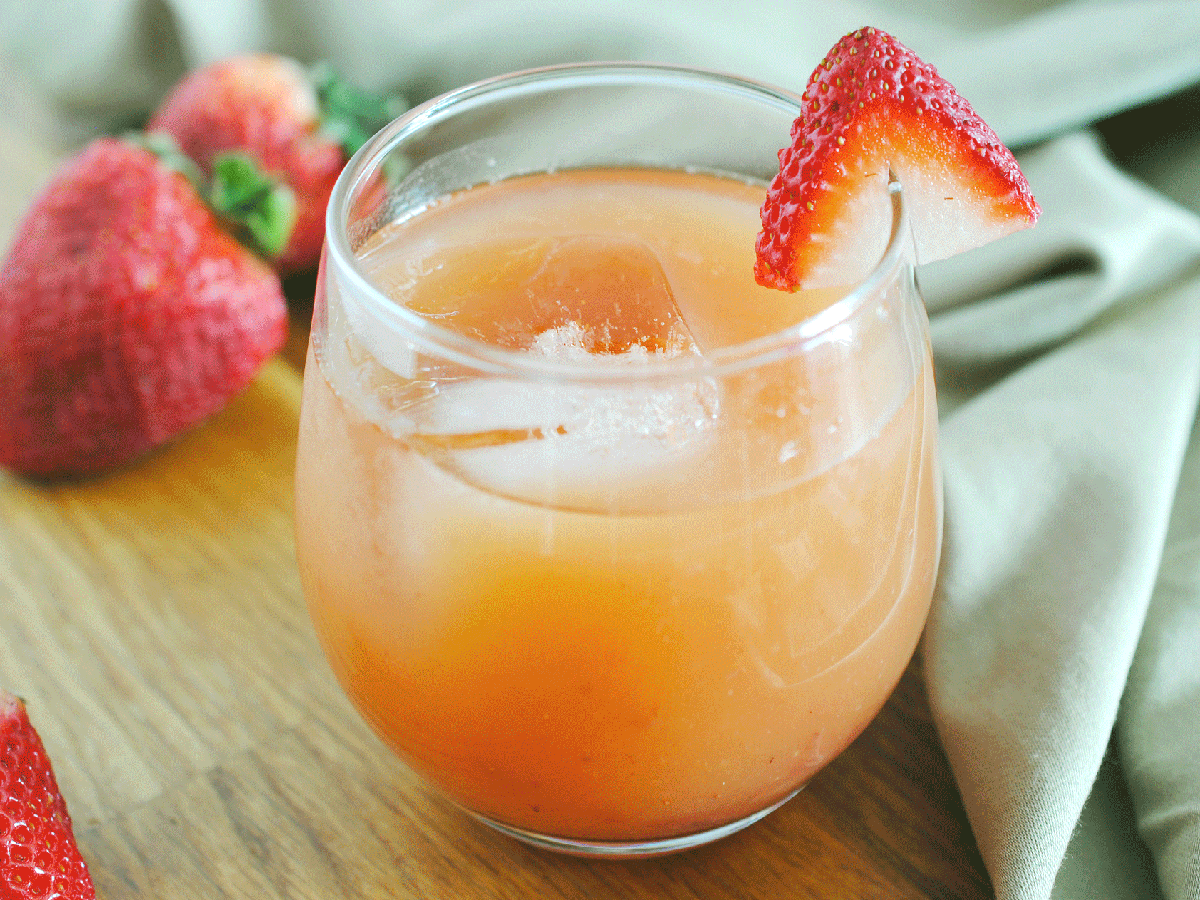 A glass of peach-flavored drink garnished with a strawberry slice, placed on a wooden surface with strawberries in the background.