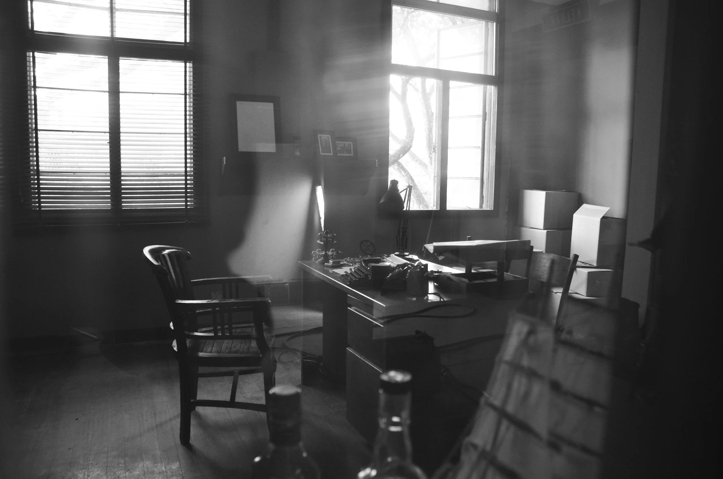 An office with a wooden desk, two chairs, and sunlight coming through the windows, creating a high contrast black and white scene.