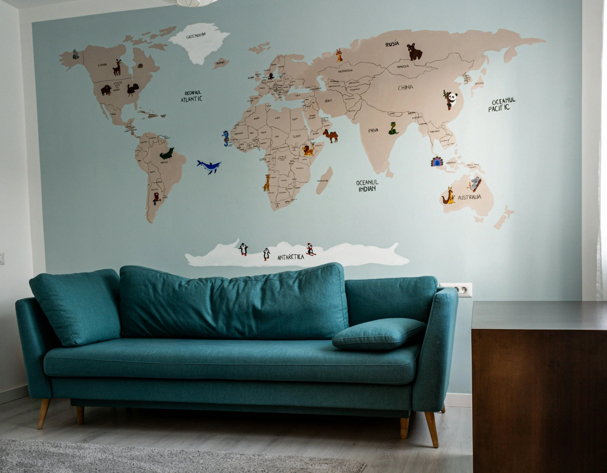 World map mural with animal illustrations of different animals in each continent, located in a room above a teal sofa.