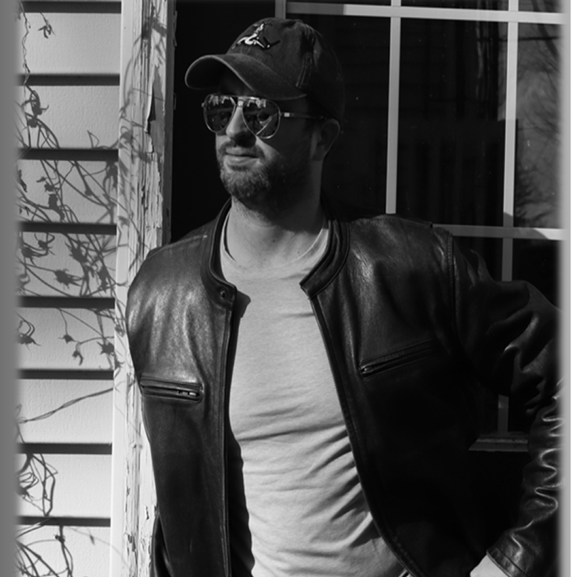 A man wearing sunglasses, a baseball cap, a leather jacket, and a T-shirt, standing outdoors near a window and some vines.