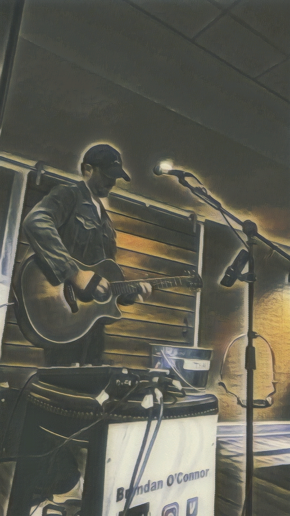 A man wearing a dark jacket and cap playing an acoustic guitar on a stage with a microphone and sound equipment