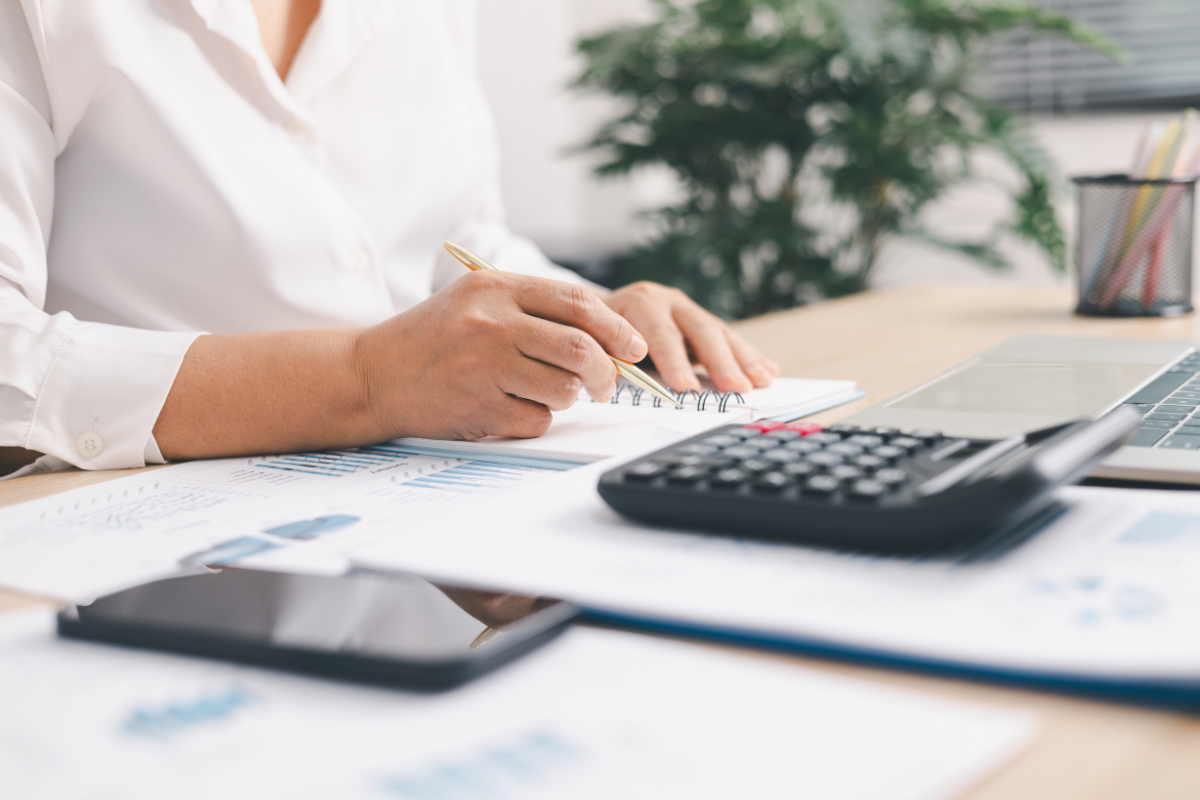 A person working at a desk with papers, a calculator, a laptop, a smartphone, and a pen, with a green plant in the background.